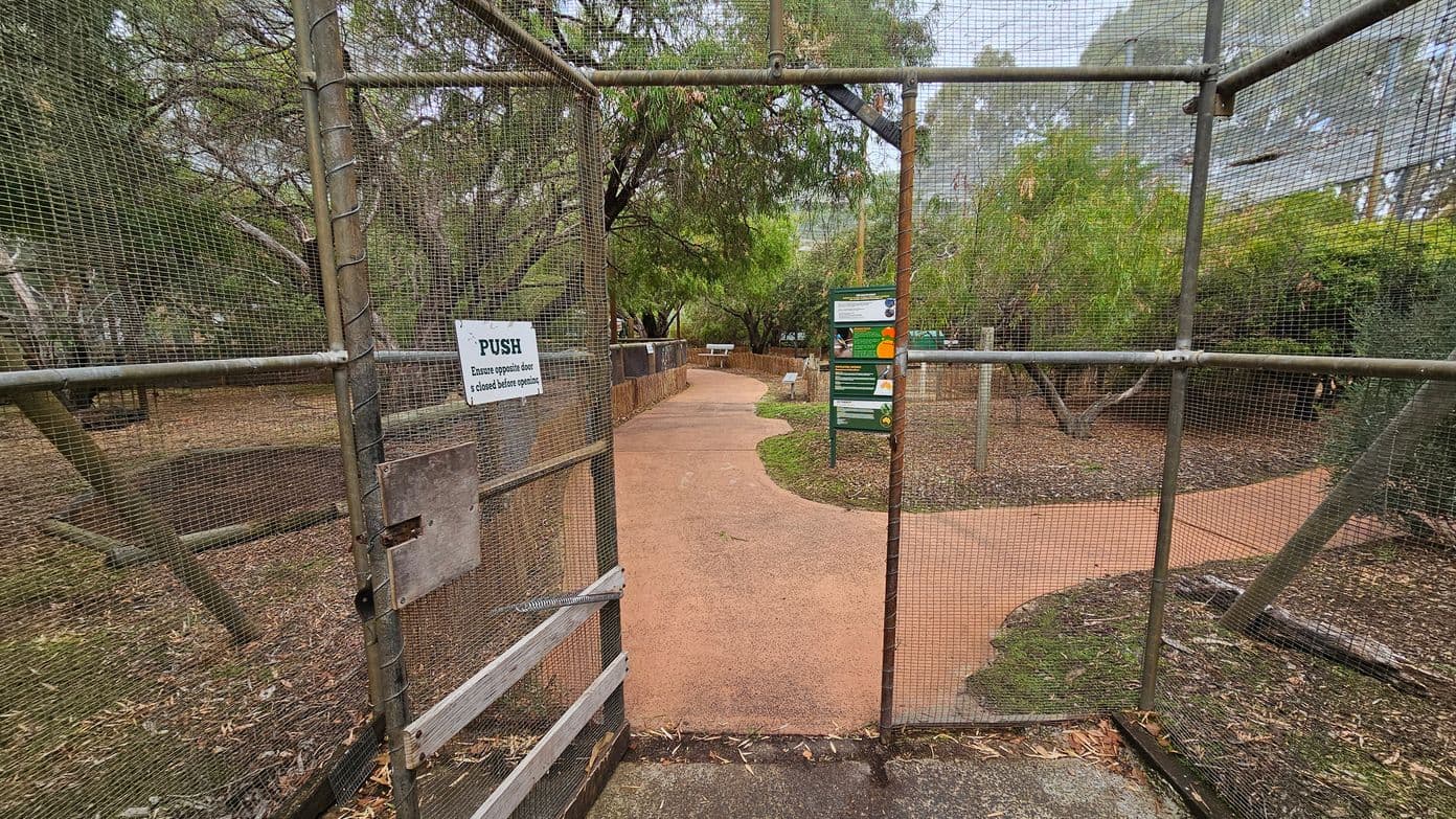 A metal mesh gate with a "PUSH" sign is in the foreground, leading to a pathway. The path curves through a garden area with trees and shrubs on both sides. In the middle distance, there is a green information board and a picnic table. The ground is covered with mulch and leaves, and the path is a reddish-brown colour. The area is surrounded by dense greenery.