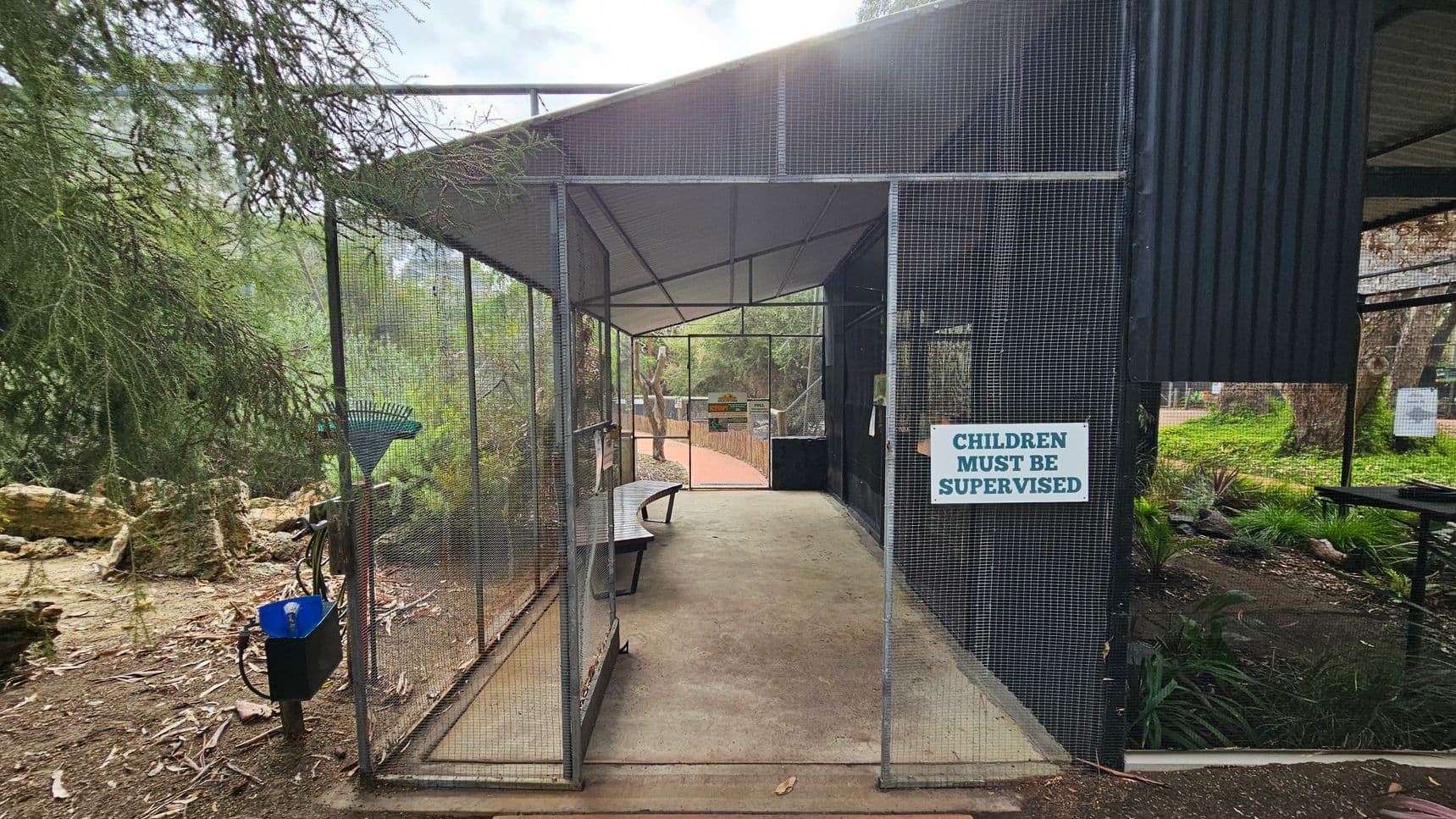 A wire mesh enclosure with a sign reading "CHILDREN MUST BE SUPERVISED" on the right. Inside, a curved bench is positioned along the left side. The path leads to a wooden fence and more greenery in the background. On the left, there are trees and a rake leaning against the mesh. The ground is covered with dirt and leaves, and the ceiling is a simple metal structure.