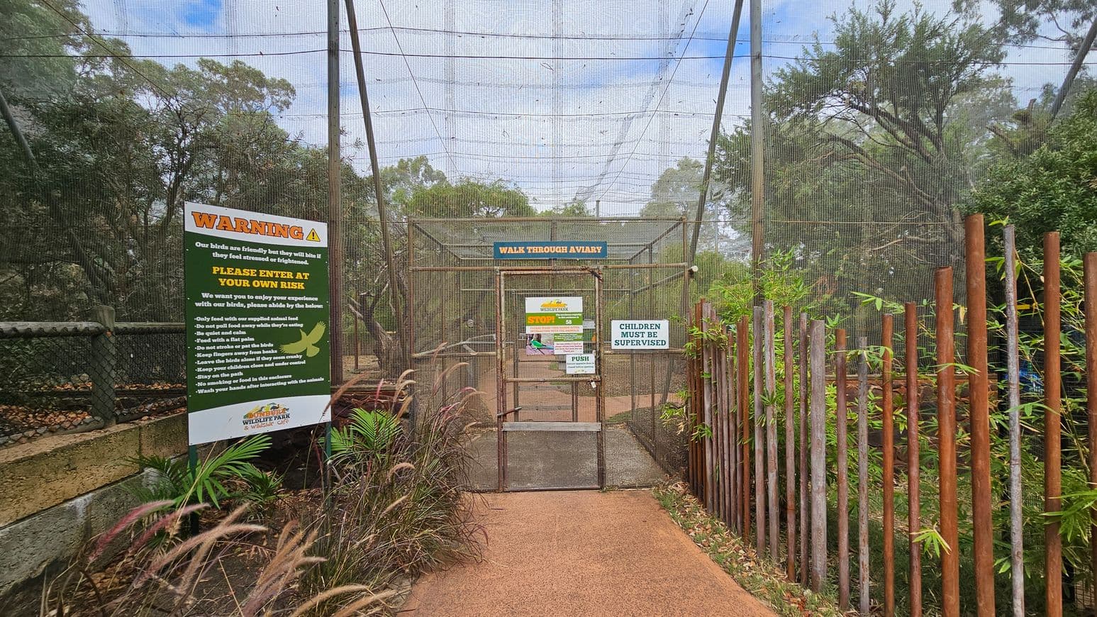 A pathway leads to a walk-through aviary entrance, enclosed with mesh netting. On the left, a large warning sign provides safety instructions. On the right, a wooden fence with vertical posts is partially covered by greenery. The aviary gate displays multiple signs, including one stating "Children Must Be Supervised." Trees and foliage are visible in the background, creating a natural setting. The ground is covered with reddish-brown gravel.