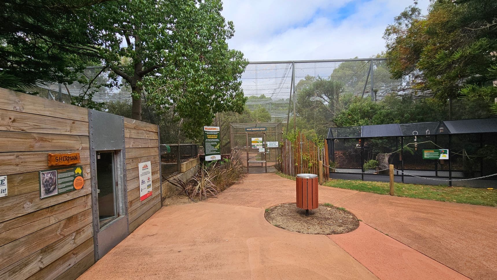 A zoo enclosure with a wooden structure on the left, featuring a sign labelled "SHERMAN" and informational posters. In the centre, a path leads to a gated area with signs, surrounded by trees and foliage. On the right, there is a black mesh enclosure with additional signs. The ground is a reddish-brown paved surface, and a circular bin is positioned in the middle of the path. The background is filled with greenery and netting above.