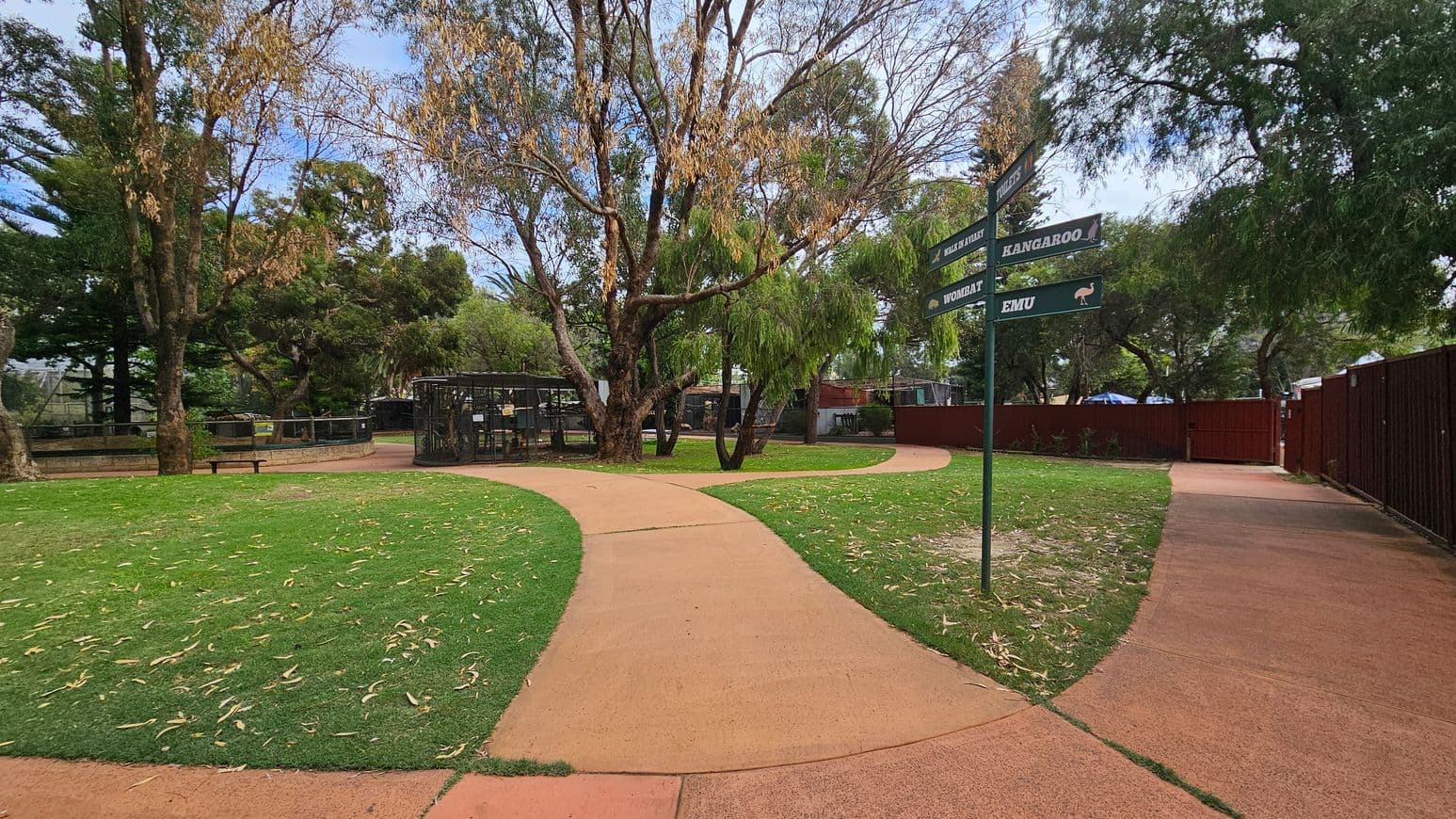 A park scene with a winding path in the centre, surrounded by green grass and scattered leaves. On the left, there are large trees and a bench near an animal enclosure. In the middle, a signpost points to various attractions like "Kangaroo" and "Emu". On the right, a red fence runs alongside the path, with more trees in the background. The area is shaded by the trees above.