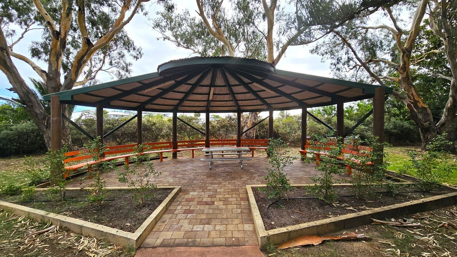 A circular gazebo with a green roof and wooden beams stands in the centre, surrounded by trees. Inside, orange benches line the perimeter, and a metal picnic table is in the middle. The floor is paved with bricks, and small plants are in raised beds at the entrance. The background features tall trees with slender trunks and leafy branches. The ground is covered with grass and scattered leaves.