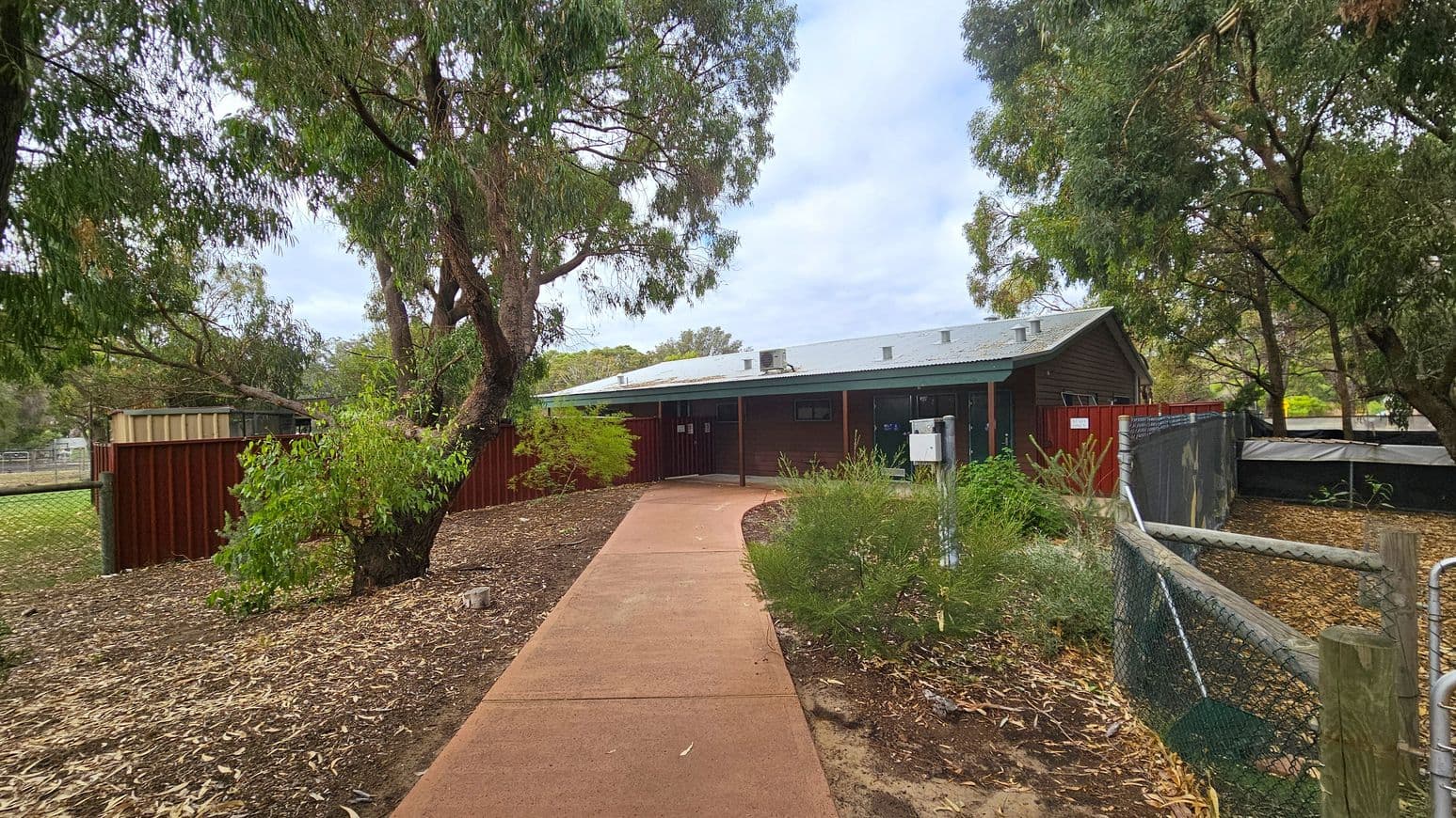 A pathway leads to a building with a grey roof and dark exterior. On the left, there are trees and a red fence, with a small shed visible. On the right, more trees and a chain-link fence with a black covering. The ground is covered with dry leaves and mulch.