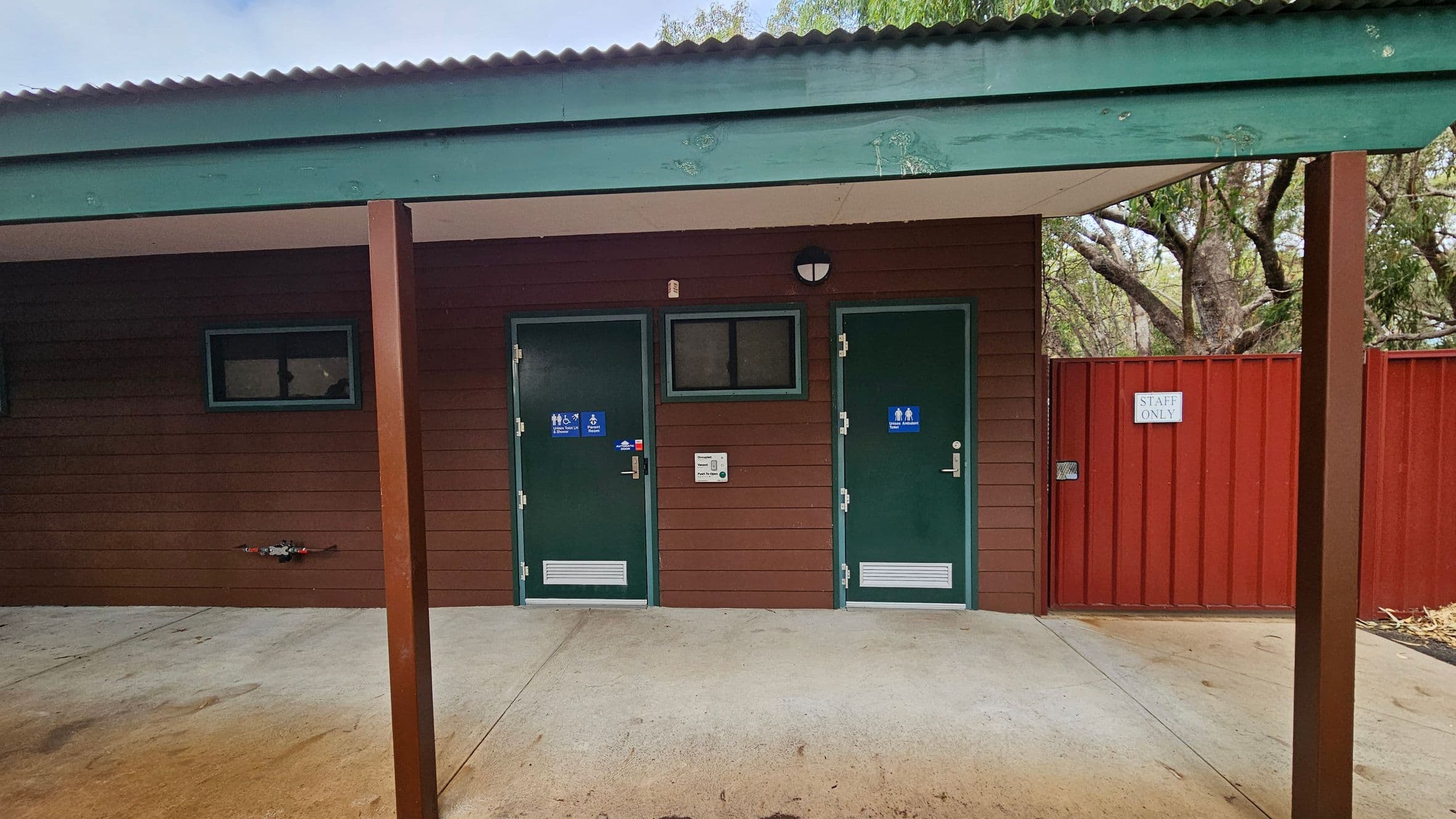 A building with a corrugated green roof and brown wooden walls. Two green doors are in the centre, each with a blue sign indicating accessible toilets. A small window is between the doors. To the right, a red fence with a "Staff Only" sign. The ground is concrete, and there are wooden posts supporting the roof. Trees are visible in the background.
