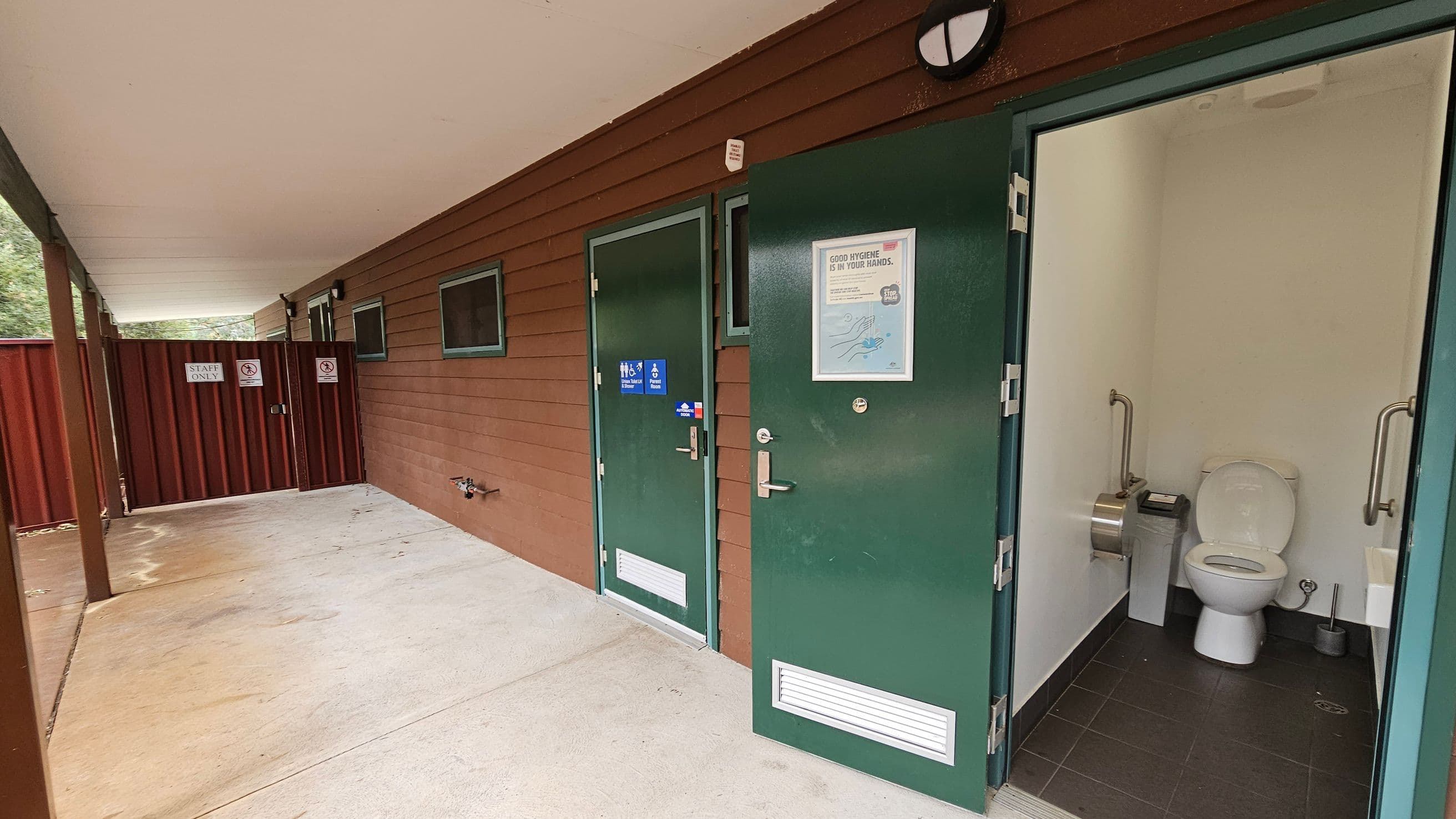 A public restroom area with a brown wooden exterior wall. On the right, a green door is open, revealing a toilet with handrails, a toilet paper dispenser, and a small sink. The floor inside is tiled in dark grey. To the left, another closed green door has a sign indicating accessibility. Further left, a red fence with "Staff Only" and no smoking signs. The ceiling is white, and the floor outside is light-coloured concrete.