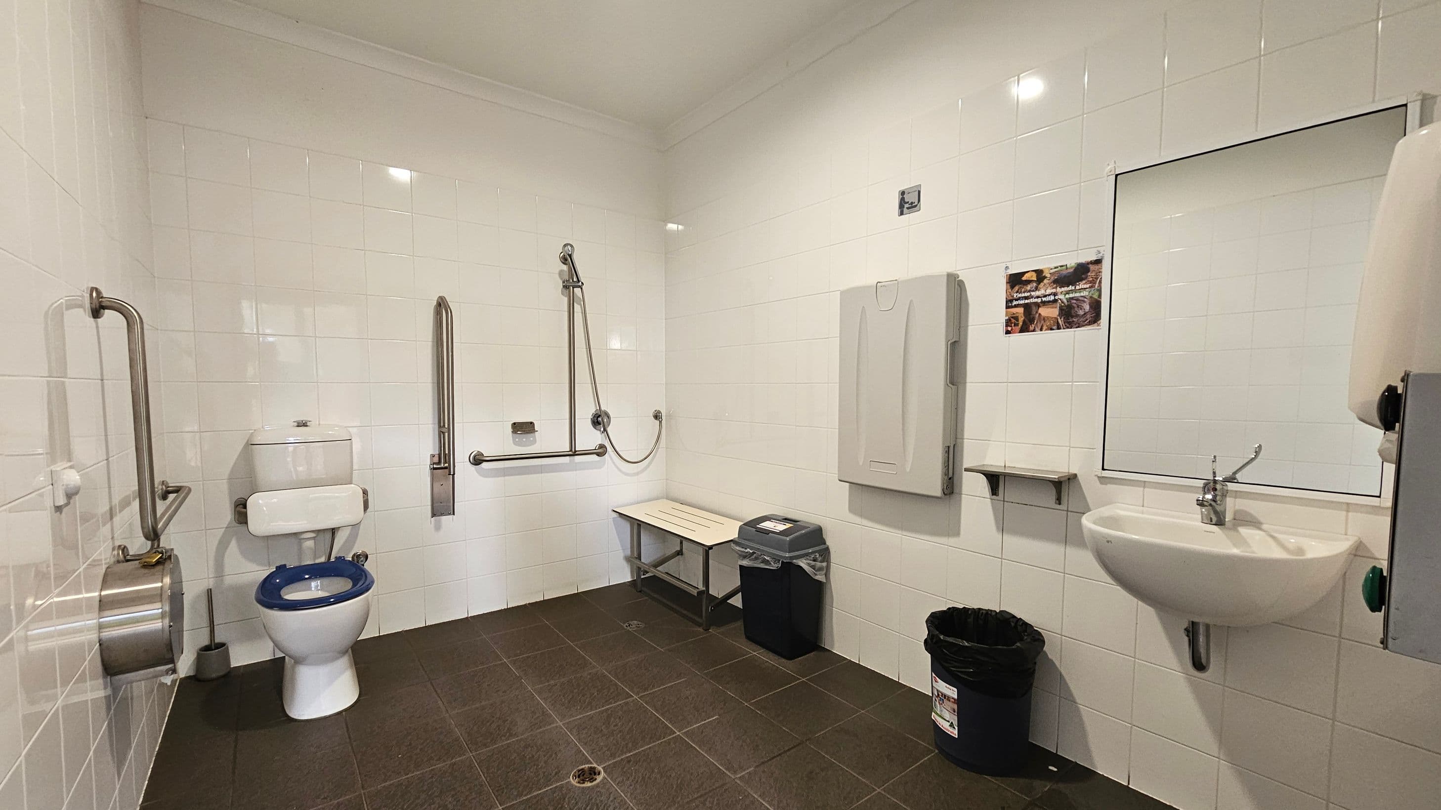 A spacious, accessible bathroom with white tiled walls and a dark tiled floor. On the left, there is a toilet with a blue seat, equipped with stainless steel grab bars. In the centre, a shower area with additional grab bars and a foldable bench. On the right, a sink with a mirror above it, a soap dispenser, and a nappy changing station. Below the mirror, a small shelf and a bin are visible. The ceiling is plain with recessed lighting.