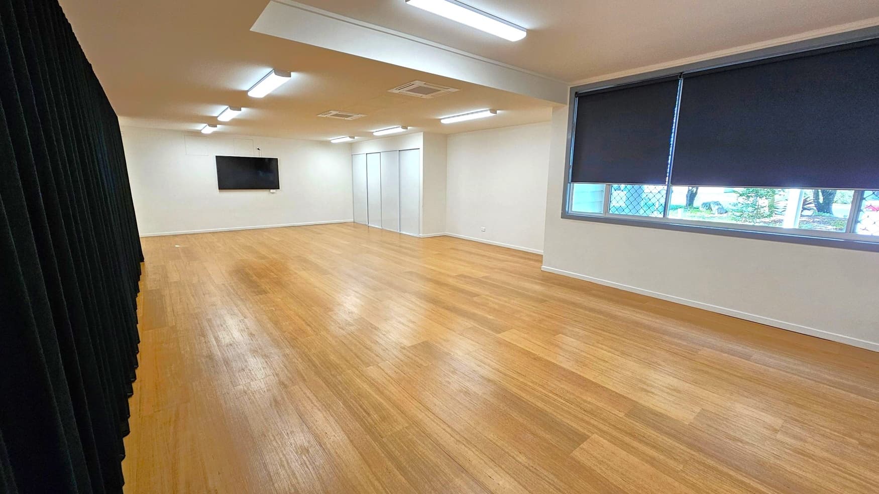 A spacious room with wooden flooring and a white ceiling featuring fluorescent lights. On the left, there is a black curtain. The right side has a large window with dark blinds partially drawn, revealing a view of trees outside. At the far end, a flat-screen TV is mounted on the wall, and there are two white doors. The room has a clean, minimalist appearance.