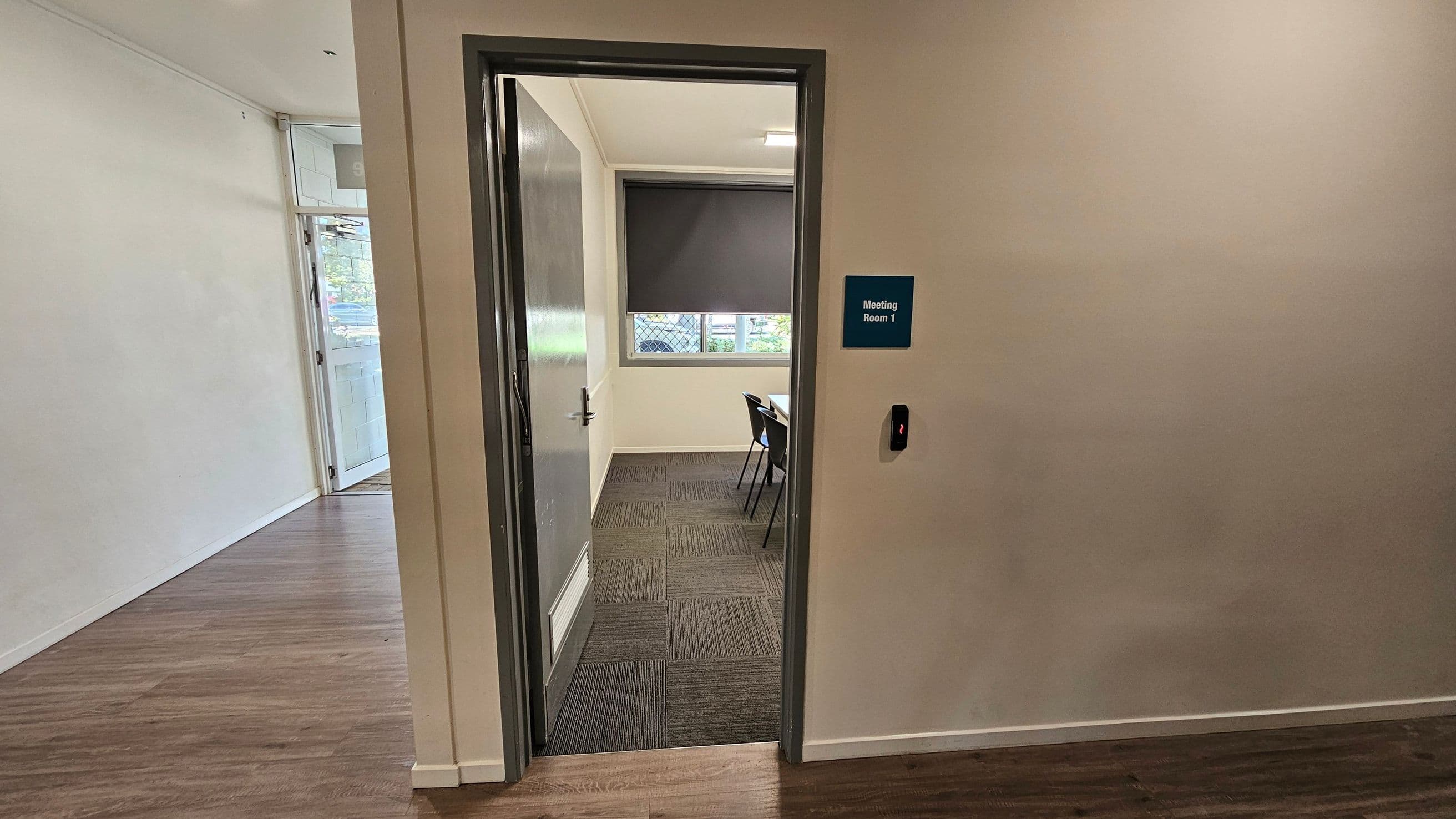 A partially open door leads into a room labelled "Meeting Room 1" on a blue sign. The room has a window with a closed blind and a table with chairs. The floor inside the room is carpeted, while the hallway has a wooden floor. A card reader is mounted on the wall next to the door. The walls are plain, and the ceiling is not visible.
