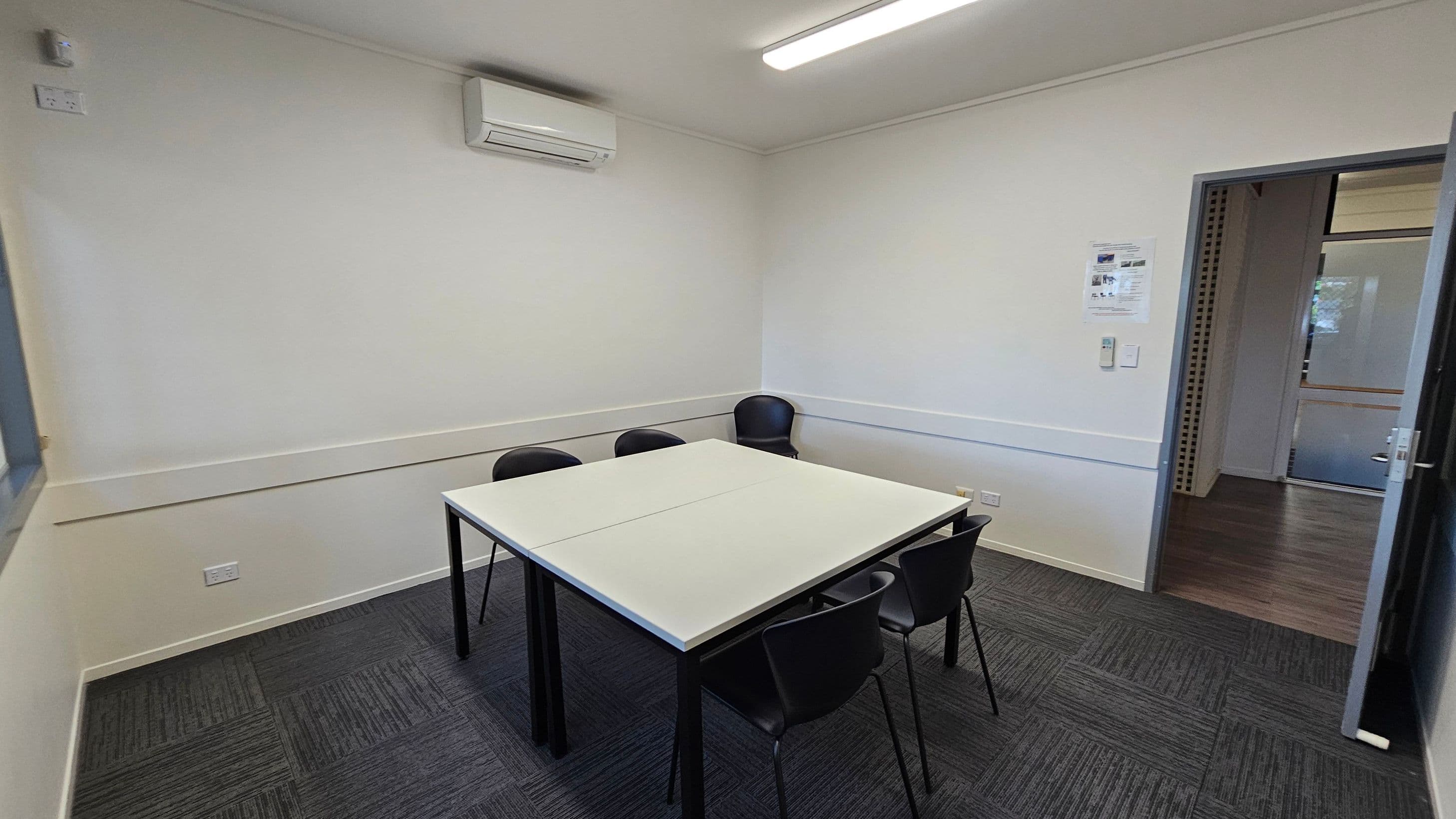 A small meeting room with a square table in the centre surrounded by four black chairs. The walls are plain white, and there's an air conditioning unit mounted on the left wall. The floor is covered with dark grey carpet tiles. On the right wall, there is a door leading to another room with wooden flooring. A fluorescent light fixture is on the ceiling, and a small notice is posted on the right wall near the door.