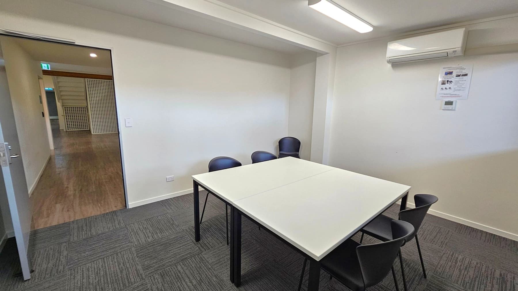 A small meeting room with a white table in the centre surrounded by four black chairs. The floor is covered with dark grey carpet tiles. On the right wall, there is an air conditioning unit and a small poster. The ceiling has a fluorescent light fixture. An open door on the left leads to a hallway with wooden flooring and a green lattice partition.