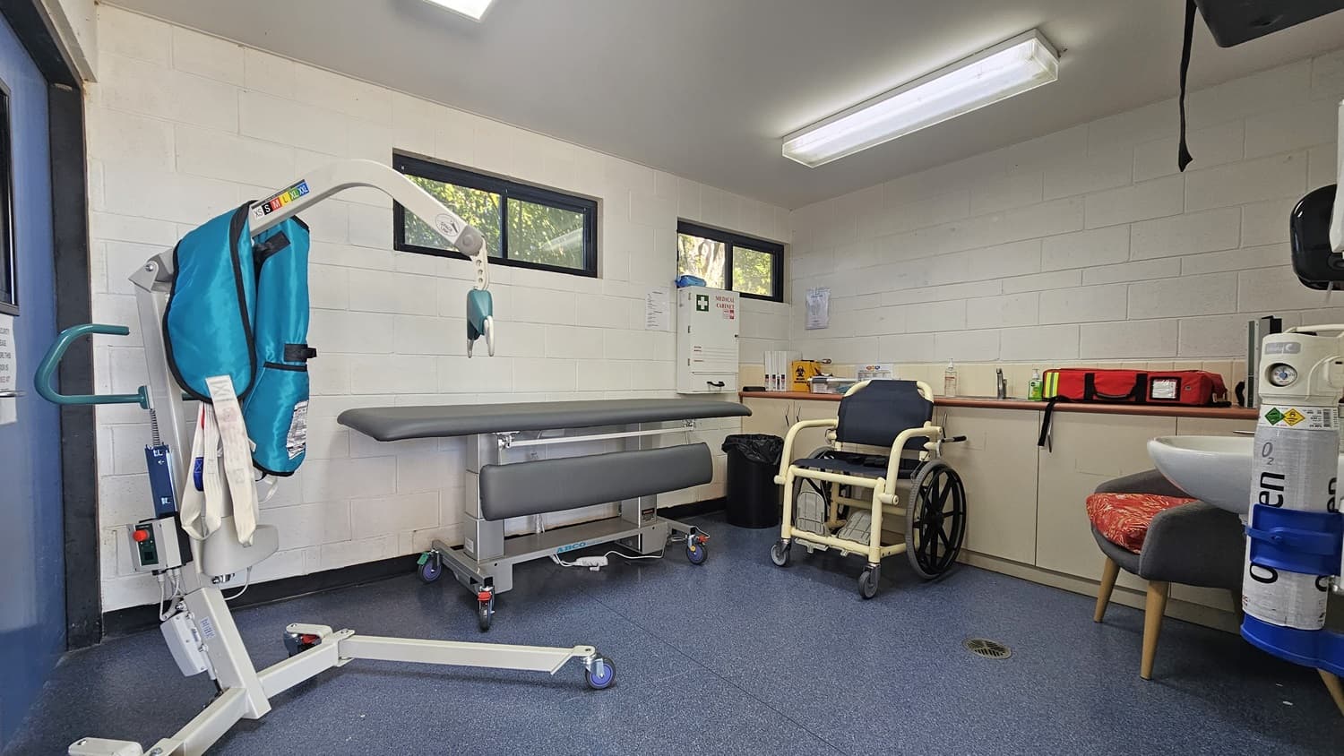 A medical room with a blue floor and white ceiling. On the left, there's a hoist with a blue sling. In the centre, a grey adjustable examination table is positioned against the wall. On the right, a wheelchair is next to a counter with medical supplies, including a red bag. A sink is visible with an oxygen cylinder nearby. The walls have small windows near the ceiling, allowing natural light in.