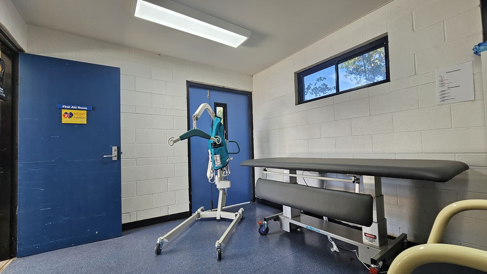 A room with white brick walls and a blue door on the left labelled "First Aid Room". In the centre, there's a mobile patient lift with a teal harness. On the right, a black cushioned examination table with metal legs is positioned against the wall. Above the table, a small rectangular window shows some trees outside. The floor is covered with blue linoleum, and a fluorescent light fixture is mounted on the ceiling.