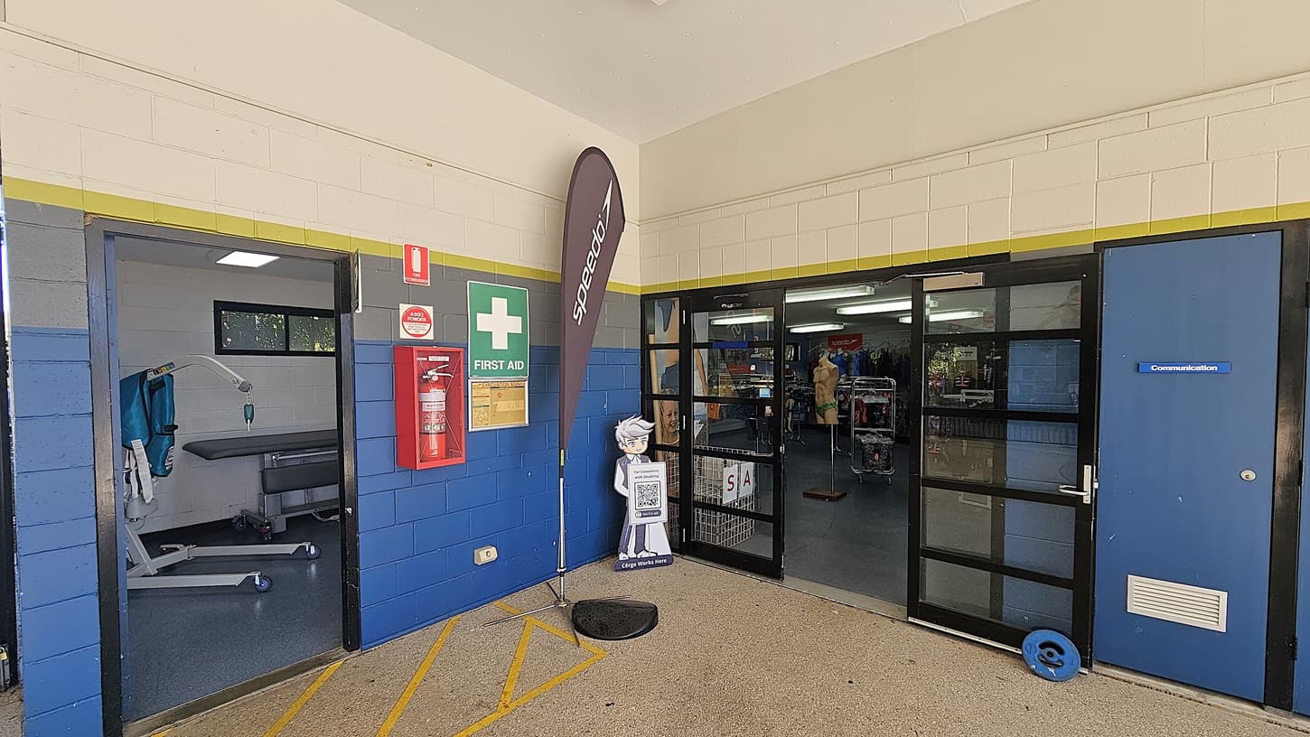 On the left, there's an open doorway leading to a room with a medical examination table and equipment. A fire extinguisher and a first aid sign are mounted on the blue and cream wall. In the centre, a vertical flag with "Speedo" written on it stands next to a cartoon figure holding a QR code sign. On the right, glass doors open into a room with visible shelves and mannequins. A blue door with a sign is adjacent to the glass doors. The floor is speckled and the ceiling is plain.
