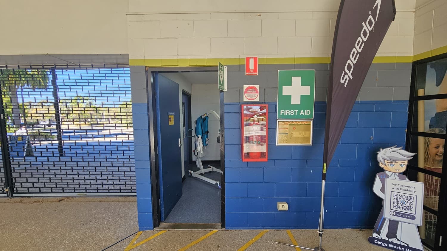 A blue and grey brick wall with a first aid sign and fire extinguisher case. An open door reveals a room with medical equipment. To the right, a vertical Speedo banner stands next to a cartoon cutout holding a QR code sign. On the left, a metal security gate shows an outdoor area with trees and a pool. The floor is concrete with yellow markings.