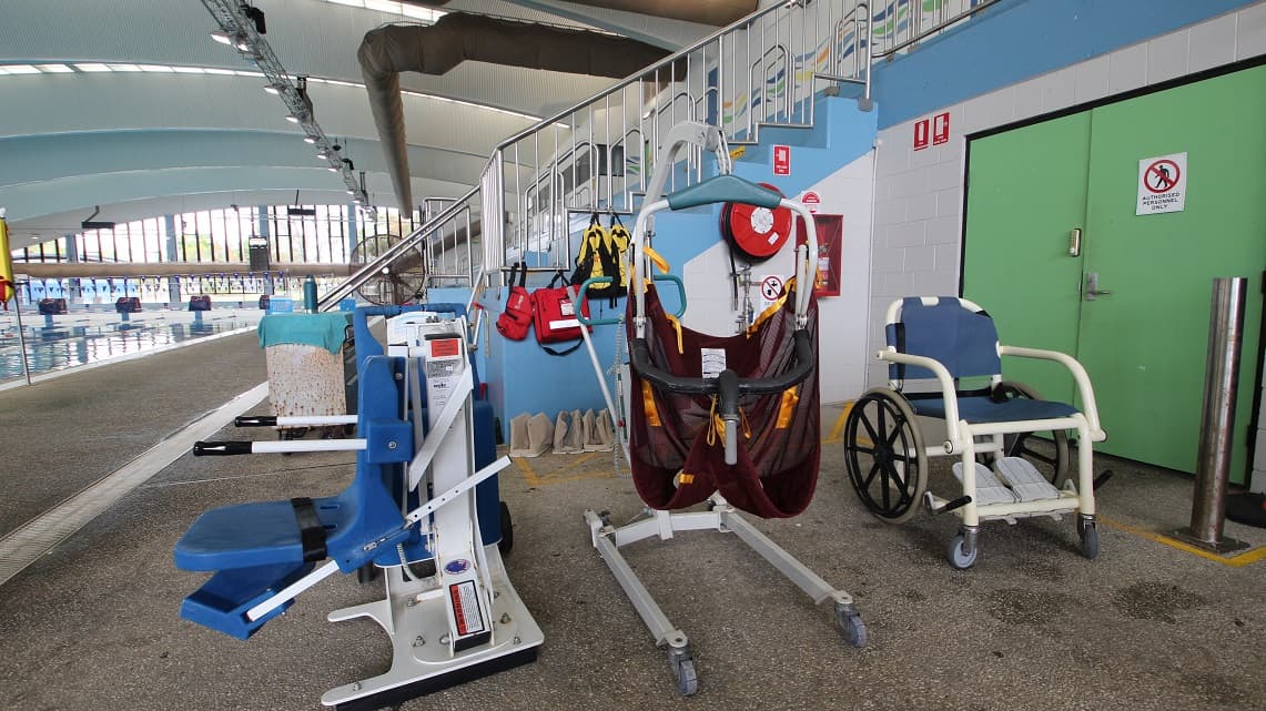 On the left, there is a blue pool lift chair. In the centre, a hoist with a maroon sling and life jackets hanging on the wall. On the right, a wheelchair with a blue seat. The background features a swimming pool with lanes and large windows. The ceiling is curved with exposed beams, and the floor is a speckled concrete surface. A staircase with a metal railing is visible behind the equipment.
