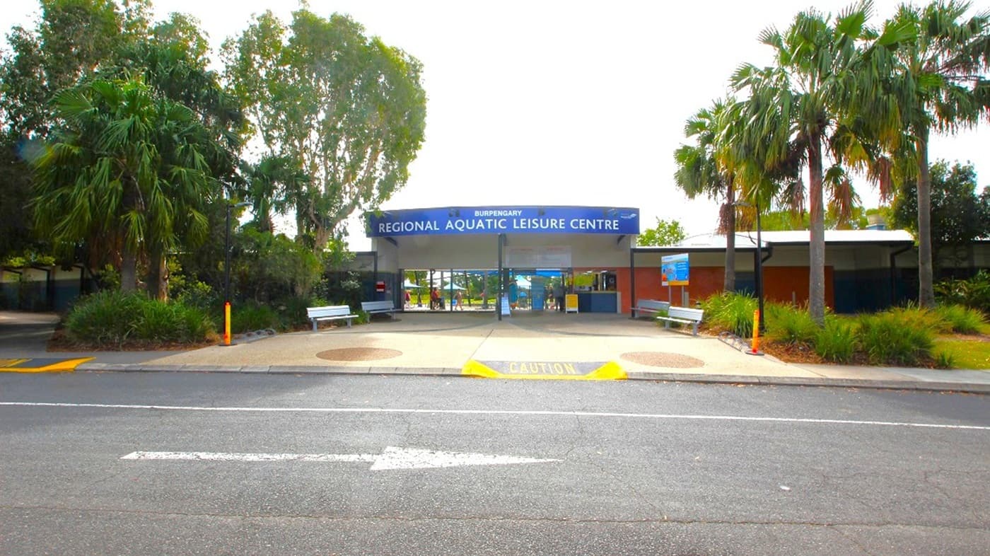 A building with a blue sign reading "Regional Aquatic Leisure Centre" is in the centre. The entrance is flanked by two white benches. On the left, there are tall trees and dense foliage. On the right, several palm trees and more greenery are visible. The foreground features a road with a pedestrian crossing marked "CAUTION" in yellow.
