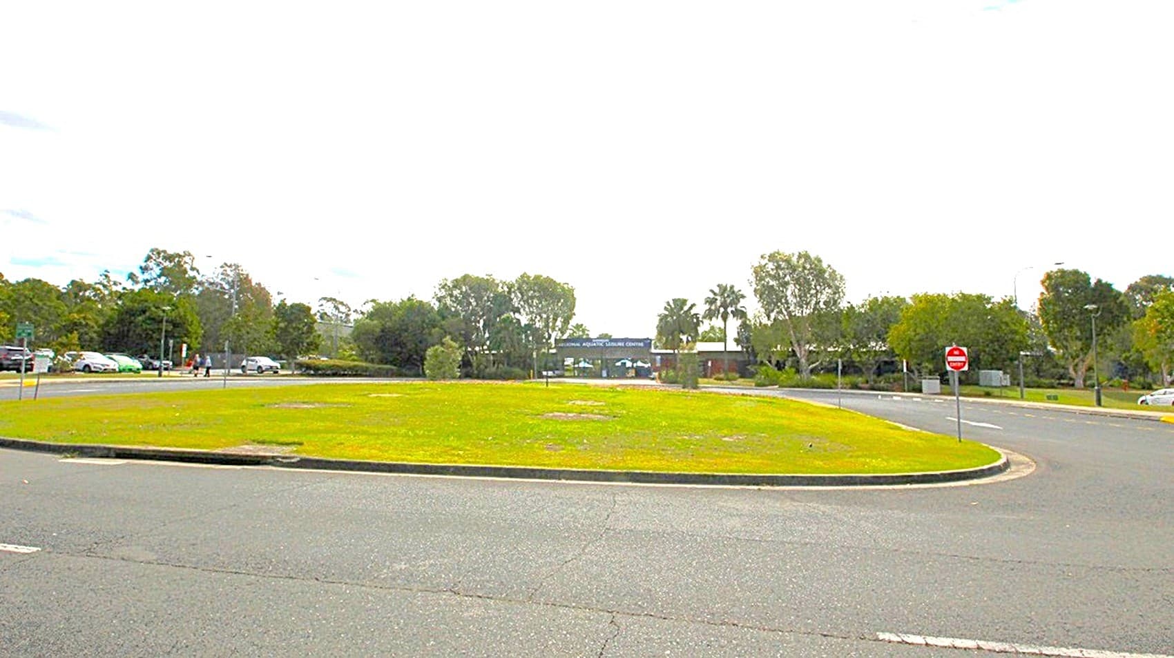 A roundabout with a grassy centre is surrounded by a paved road. On the left, there are several parked cars and trees in the background. To the right, more trees and a "no entry" sign are visible. The background features a line of trees and some buildings partially obscured by foliage. The sky is overcast with scattered clouds.
