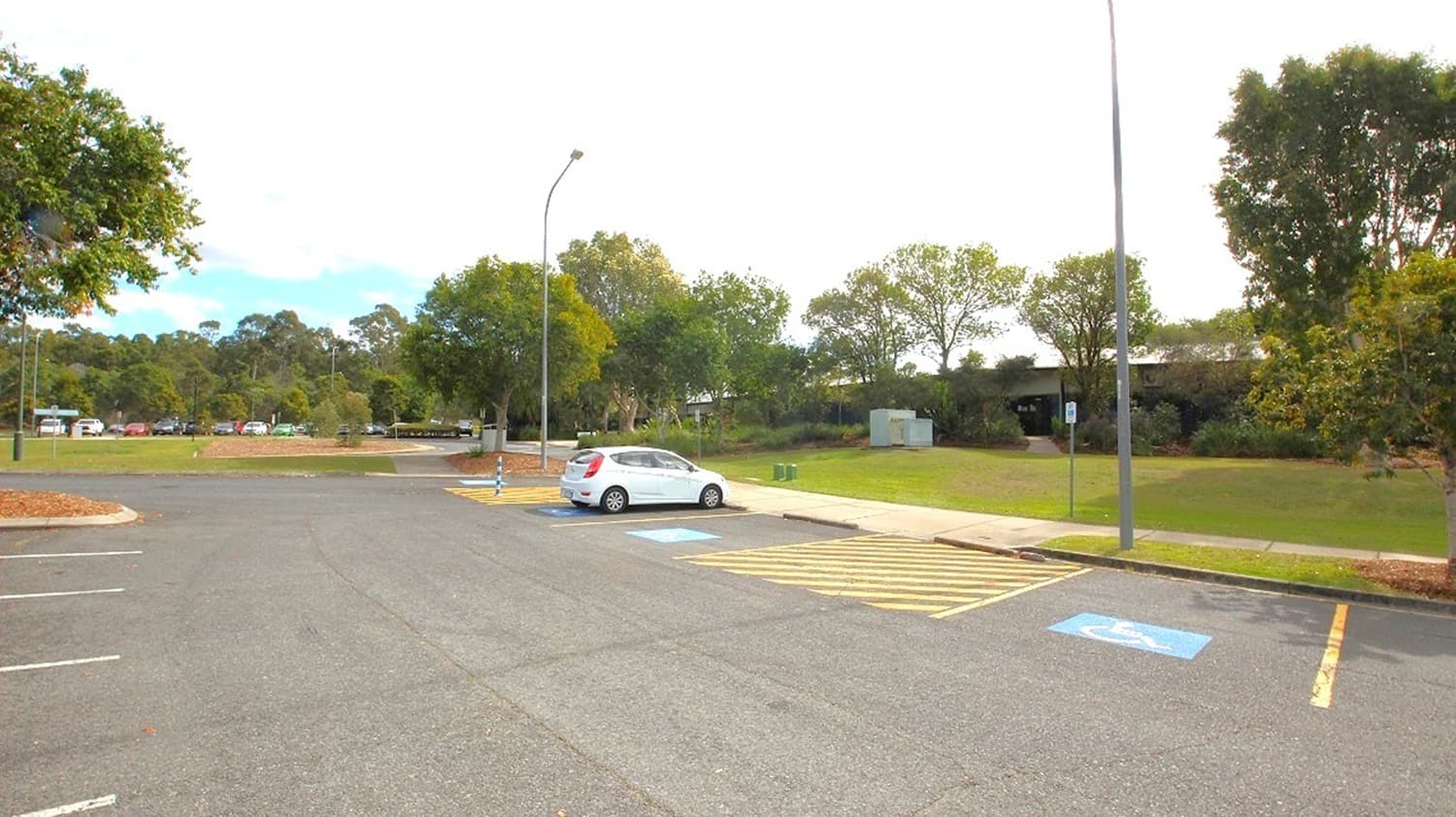 A nearly empty car park with a single white car parked in a designated disabled parking space on the right. The ground is paved with marked parking lines. Two tall streetlights are visible, one near the car and another further back. In the background, there are green trees and a grassy area. A few more cars are parked in the distance to the left, and the sky is partly cloudy.