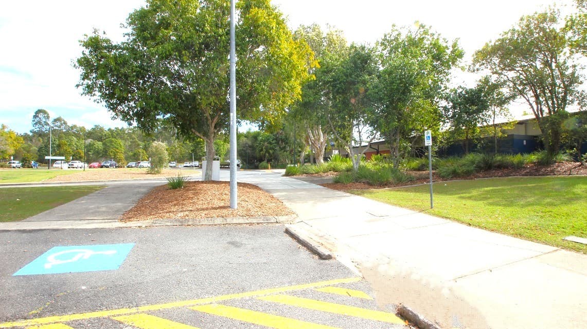 A parking area with a designated disabled parking space marked in blue on the left. A concrete path runs through the centre, bordered by a grassy area on the right. Several trees with dense foliage are scattered throughout, providing shade. A lamppost stands near the centre of the image. In the background, more trees and a few parked cars are visible, along with a building partially obscured by the greenery.