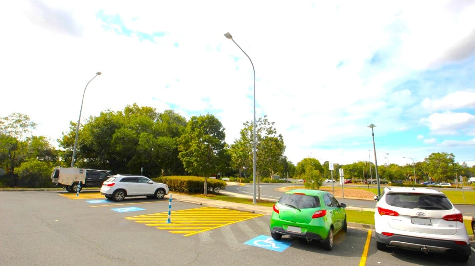 A parking lot scene with several cars parked near handicapped spaces. In the foreground, a green hatchback and a white SUV are parked in standard spots, while a white SUV and a light-colored van are parked along the curb. Blue handicapped parking signs and yellow striped crosshatched area are visible near the spaces. In the background, there are trees, a curved streetlamp, and a partly cloudy blue sky.