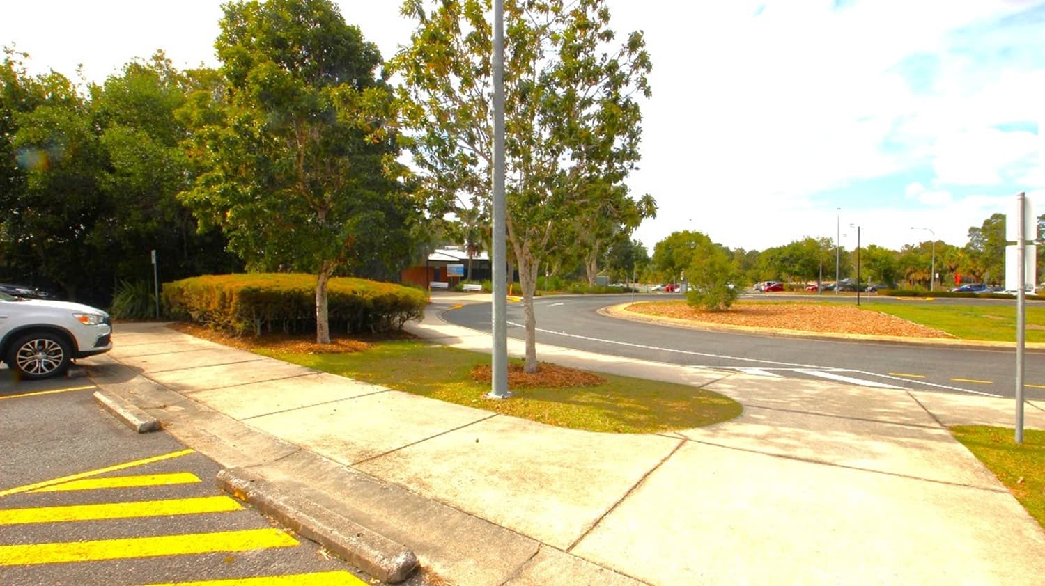 A parking lot and sidewalk scene with a curved road in the background. In the foreground, yellow striped handicap markings and a curbside curb are visible, along with a white SUV parked to the left. A sidewalk runs parallel to the curb, and a tall street lamp stands near a small island with trees and shrubs. The sky is partly cloudy with blue patches.