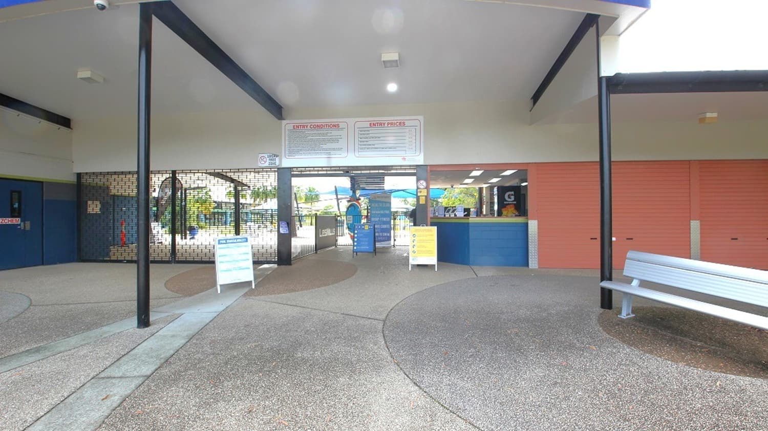 An entrance area with a covered ceiling supported by black poles. In the centre, there are signs above a gated entryway, with a view of outdoor seating and umbrellas beyond. To the right, a blue counter is visible, possibly for ticketing or information, with a brick wall behind it. A silver bench is positioned on the far right. The floor is a textured grey surface, and there are informational signs on stands near the entrance.