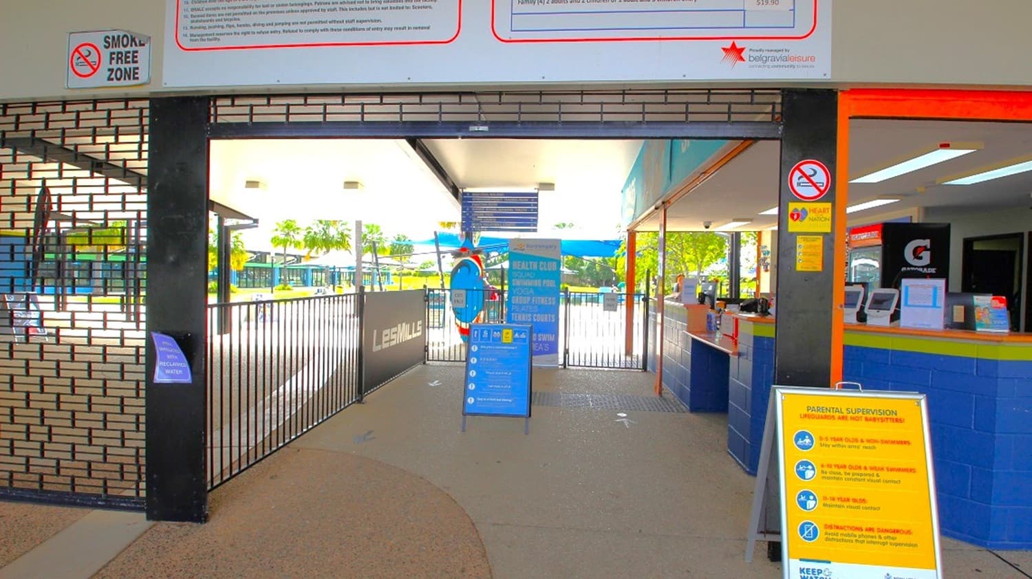 A gated entrance with a "Smoke Free Zone" sign above. On the right, there's a counter with a "G" logo and informational signs. In the centre, a blue sign with instructions stands near the gate. Beyond the gate, outdoor seating and palm trees are visible. The floor is concrete, and the ceiling is white with lights. A yellow sign with safety instructions is on the right side near the entrance.