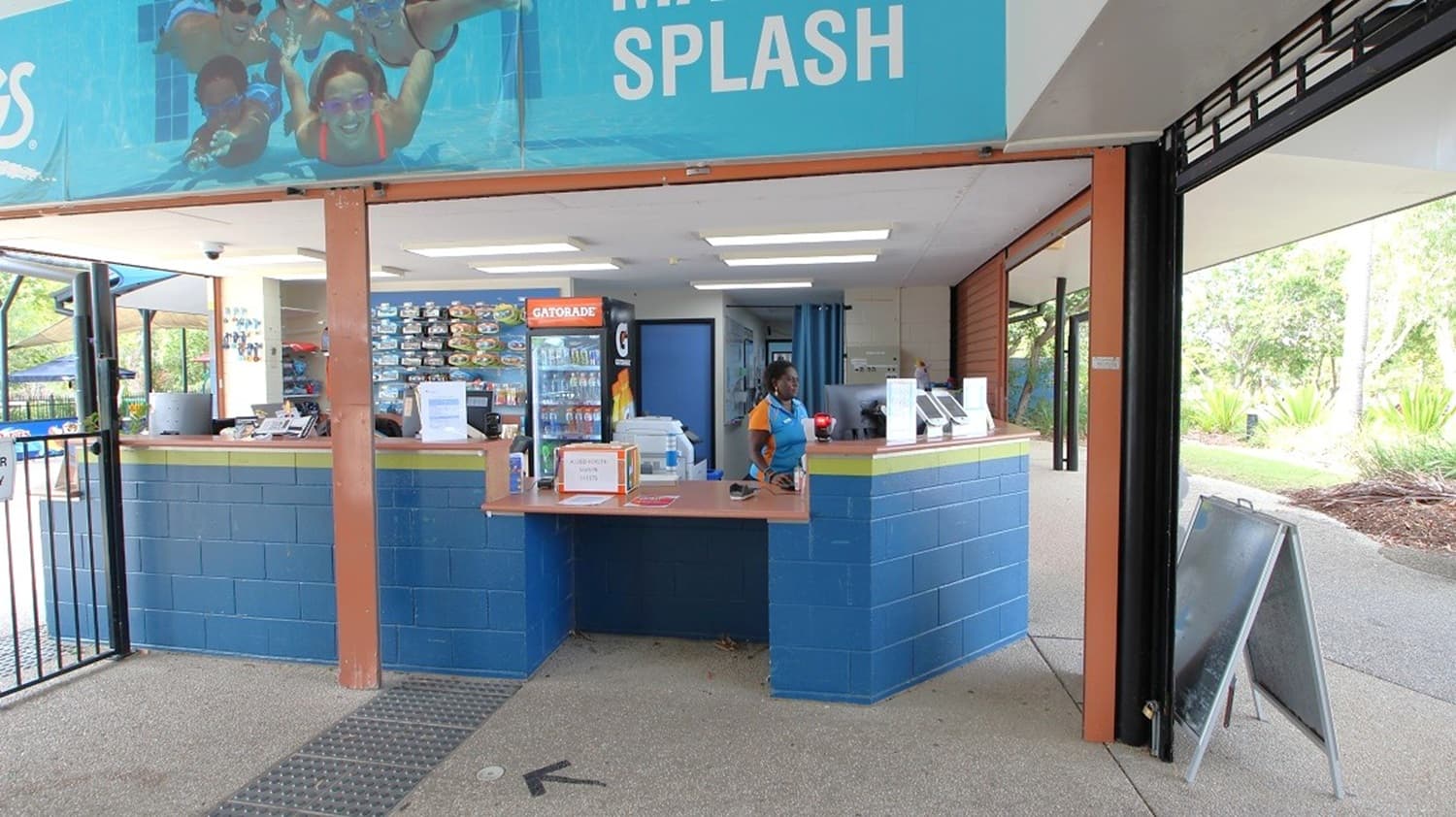 A reception area with a blue counter in the centre, staffed by a person wearing a blue and orange uniform. Behind the counter, a fridge stocked with drinks and a menu board are visible. Above, a large banner displays people enjoying a water slide. The floor is concrete, and the ceiling has fluorescent lights. On the right, a pathway leads outside, bordered by greenery. A blackboard sign stands near the entrance.