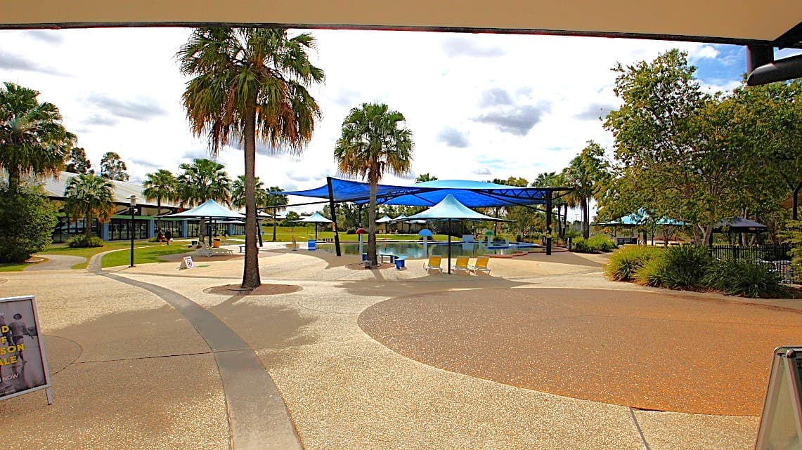 A spacious outdoor area with a curved pathway and textured ground. In the centre, there are several blue shade structures and a few yellow chairs. Palm trees are scattered throughout, providing a tropical feel. On the left, a building with large windows is partially visible. On the right, there are more trees and greenery. The sky is cloudy, and the area appears to be part of a recreational or park setting.