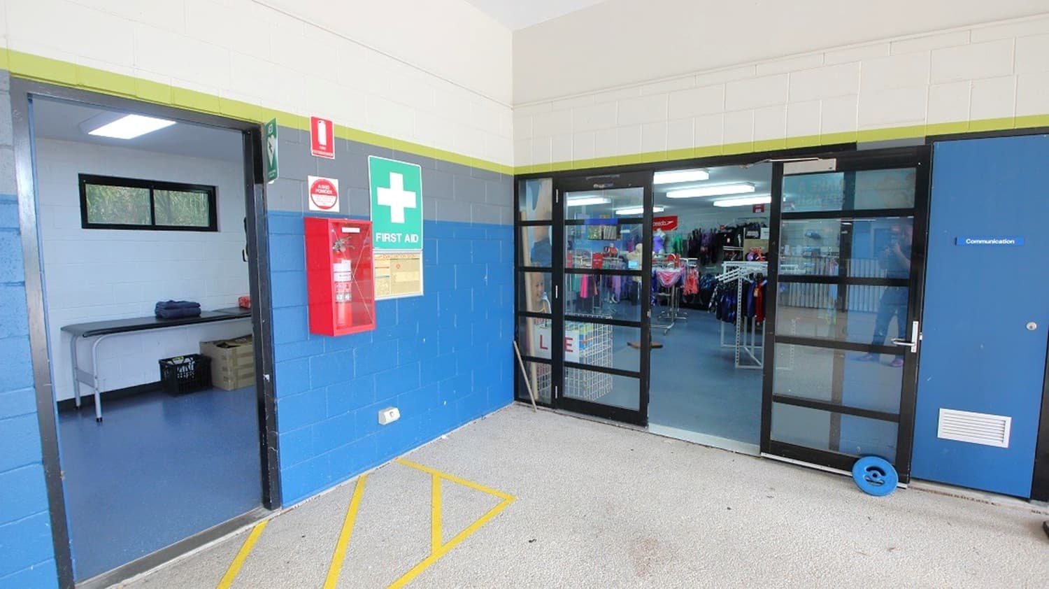 On the left, an open door leads to a room with a table and boxes. A red fire extinguisher and a first aid sign are mounted on the blue wall. In the middle, a glass door reveals a shop interior with clothing racks. On the right, a closed blue door is visible. The floor is grey with yellow markings, and the ceiling is white.