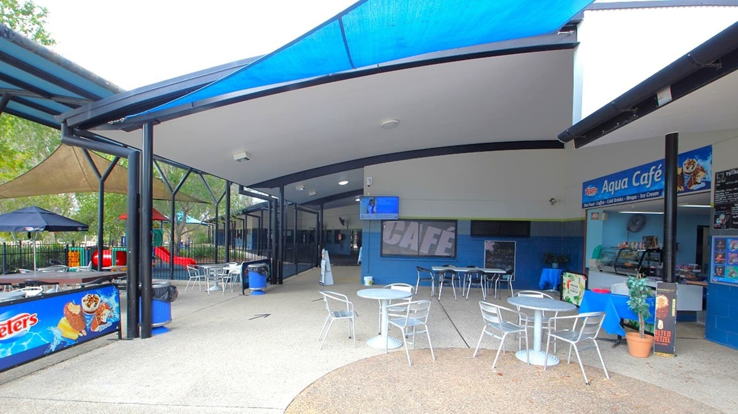 Outdoor café area with a blue and white theme. On the right, there's a counter labelled "Aqua Café" with a menu and ice cream advertisements. Several metal tables and chairs are scattered around. The left side has a shaded play area with colourful equipment and umbrellas. The ceiling is covered with blue fabric, and the floor is a mix of concrete and textured surfaces. A small potted plant is near the café counter.