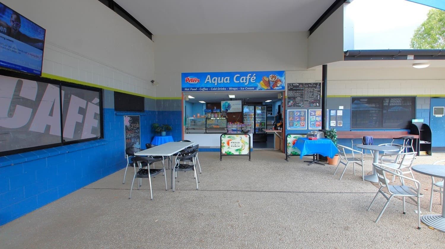 A café with a blue and white sign reading "Aqua Café" is in the centre. The entrance has a counter displaying food items. On the left, there are tables and chairs with a large sign on the wall. On the right, more tables and chairs are set up, with a menu board and a table covered with a blue cloth. The floor is a light-coloured textured surface, and the ceiling is high with exposed beams.