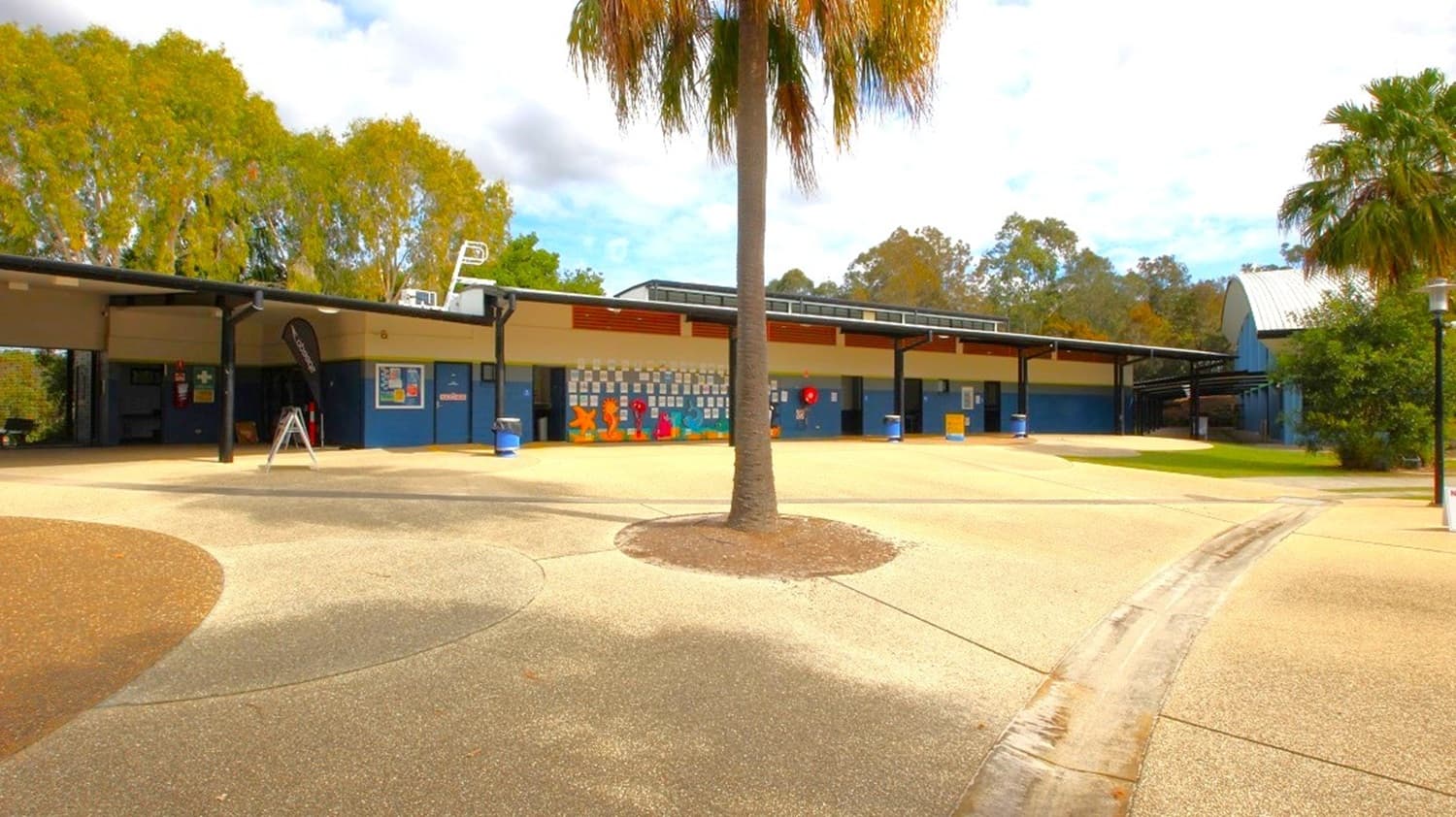 A courtyard with a central palm tree on a circular patch of soil. The ground is paved with a mix of light and dark concrete. In the background, a long, low building with a flat roof features colourful murals and several blue bins. Trees with dense foliage are visible behind the building. On the right, another palm tree and a curved path lead towards more greenery. The sky is partly cloudy.