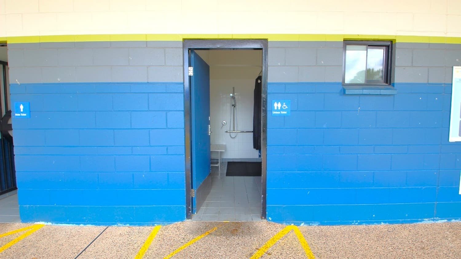 A building with a blue and grey exterior wall featuring a central open door leading to a bathroom. The bathroom has a shower and a toilet with grab bars, indicating accessibility features. A small window is on the right side of the wall. The floor outside is concrete with yellow painted lines. Signs on the wall indicate the bathroom is accessible.
