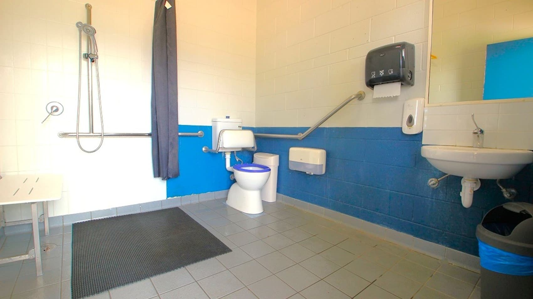 A spacious, accessible bathroom with a grey tiled floor. On the left, there's a shower area with a curtain and a grab bar. In the centre, a toilet with a blue seat is equipped with support rails. On the right, a white sink is mounted on the wall with a mirror above it. A black paper towel dispenser is also mounted on the wall. The walls are painted white with a blue accent strip running horizontally. A black mat is placed on the floor near the shower.