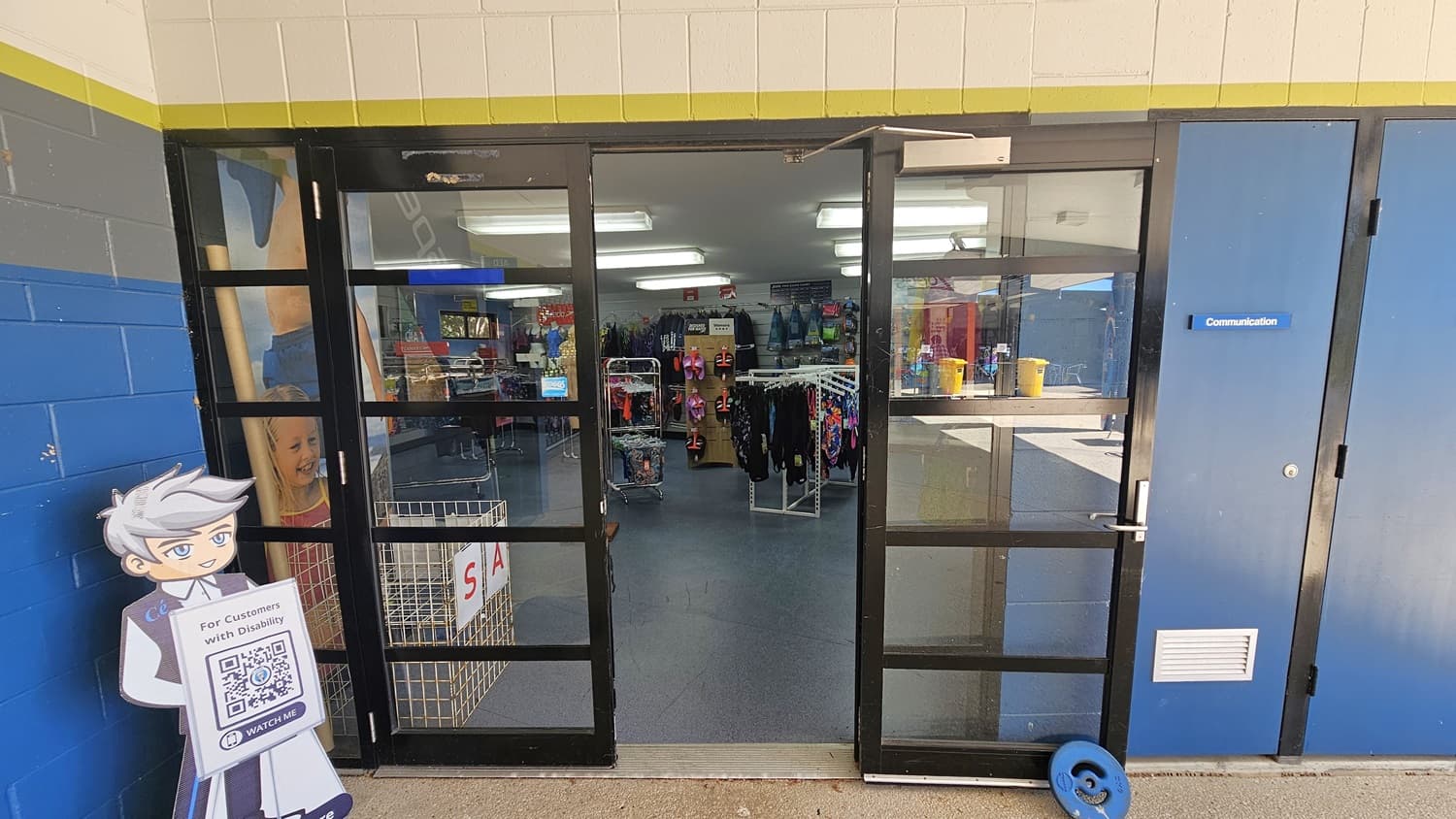 On the left, there is a life-sized cutout of a cartoon character holding a QR code sign next to a glass door. On the right, a blue door labelled "Communications" is adjacent to the glass door. In the middle, the glass doors open into a shop with clothing racks displaying various items. The background includes more clothing racks and shelves. The foreground features a concrete floor outside the shop and a blue floor inside. The ceiling inside has fluorescent lights.