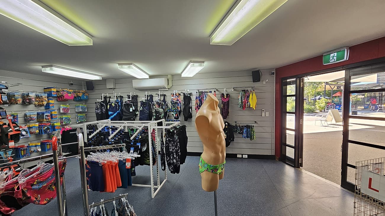 On the left, there are shelves displaying swim goggles, caps, and colourful bags, with a clothing rack holding swimwear. On the right, a glass door with an exit sign leads outside to a patio area with tables and chairs. In the middle, a mannequin torso in swim trunks stands near more racks of swimwear. The background includes additional shelves and racks with swim gear. The floor is covered with blue carpet tiles. The ceiling has rectangular fluorescent lights.