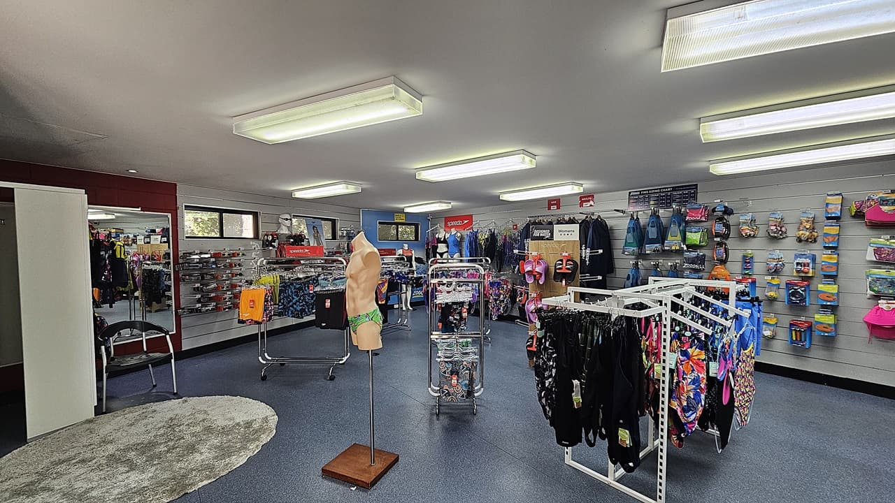 On the left, there is a white partition next to a wall with various items hanging, including bags and accessories. On the right, there are shelves displaying swimwear and colourful bags. In the middle, a mannequin torso stands on a wooden base near clothing racks filled with swimwear. The background includes more shelves with swim gear and accessories. The foreground features a blue carpeted floor with a circular grey rug. The ceiling has rectangular fluorescent lights.