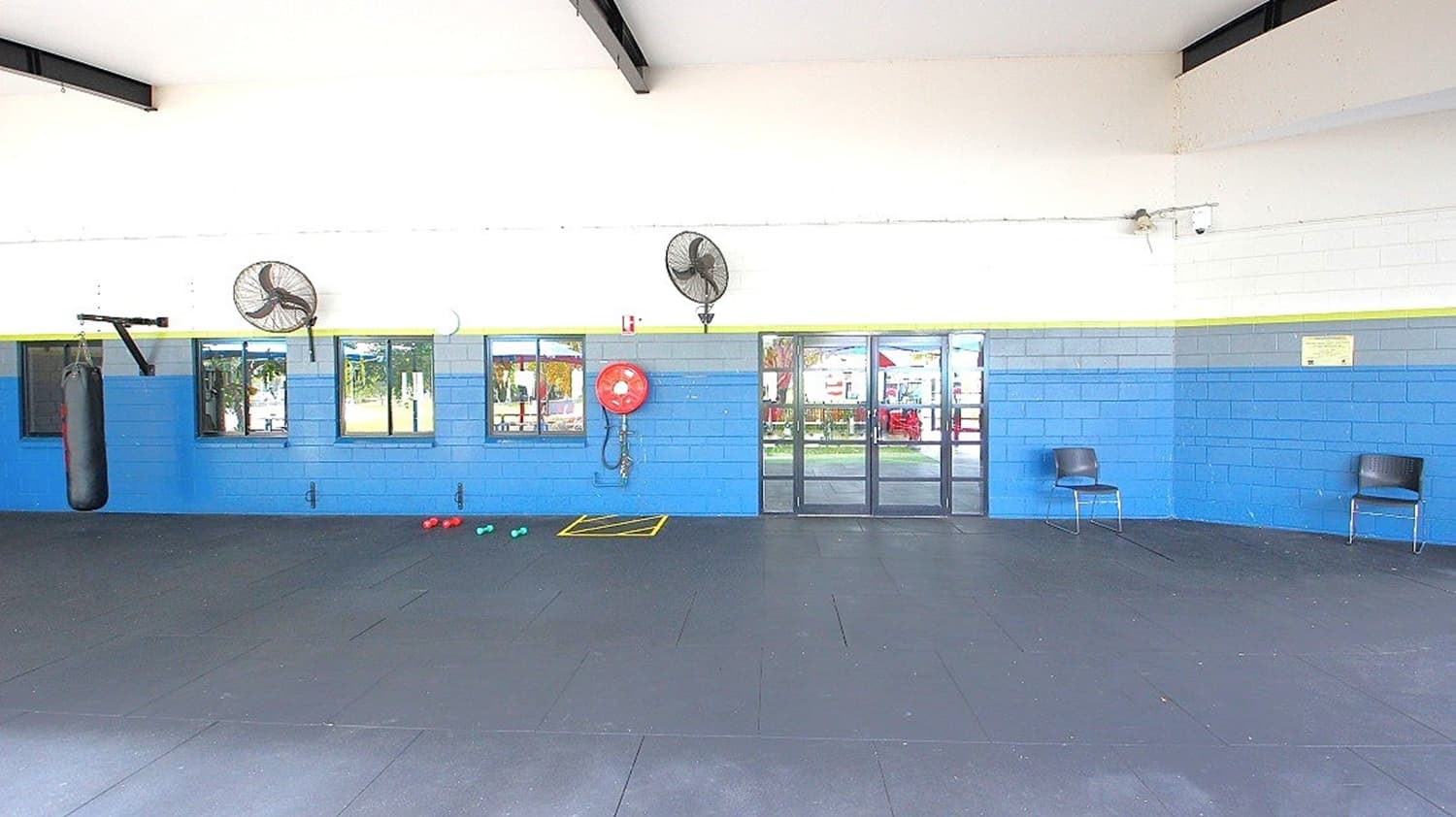 A gym interior with a blue and white colour scheme. On the left, a punching bag hangs from the wall. Three windows are visible, with two wall-mounted fans above them. In the centre, a red fire hose reel is mounted on the wall, and a set of glass doors leads outside. On the floor, there are several small balls and a yellow agility ladder. Two chairs are placed against the wall on the right. The ceiling is white with exposed black beams, and the floor is dark grey.