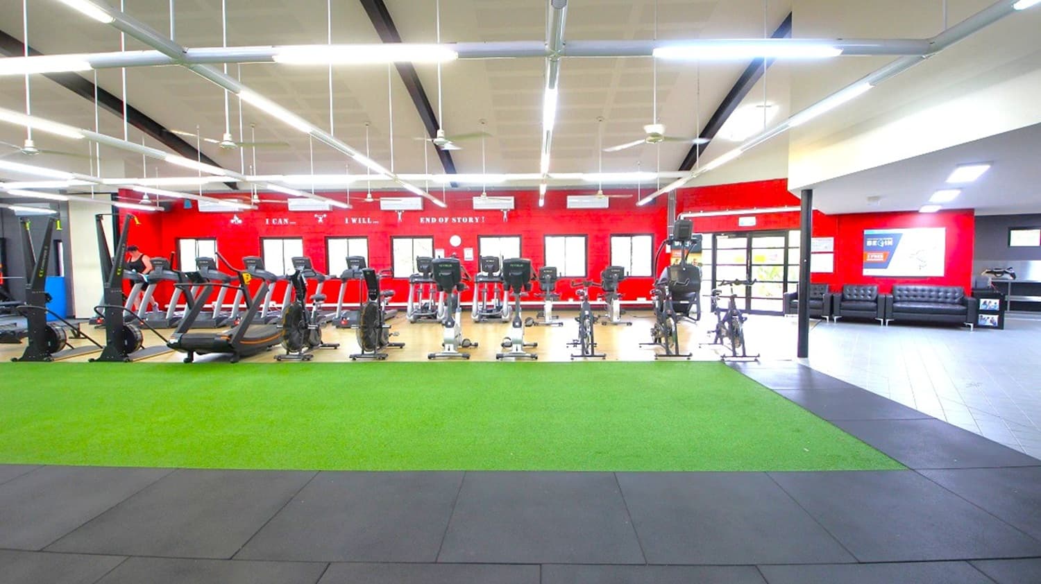 A spacious gym interior with a row of treadmills and exercise bikes lined up against a red wall in the background. The wall features motivational text and several windows. In the foreground, there is a green artificial turf area, and the floor is a mix of black and grey tiles. To the right, there is a seating area with black sofas and a wall with posters. The ceiling is equipped with bright fluorescent lights and ceiling fans.