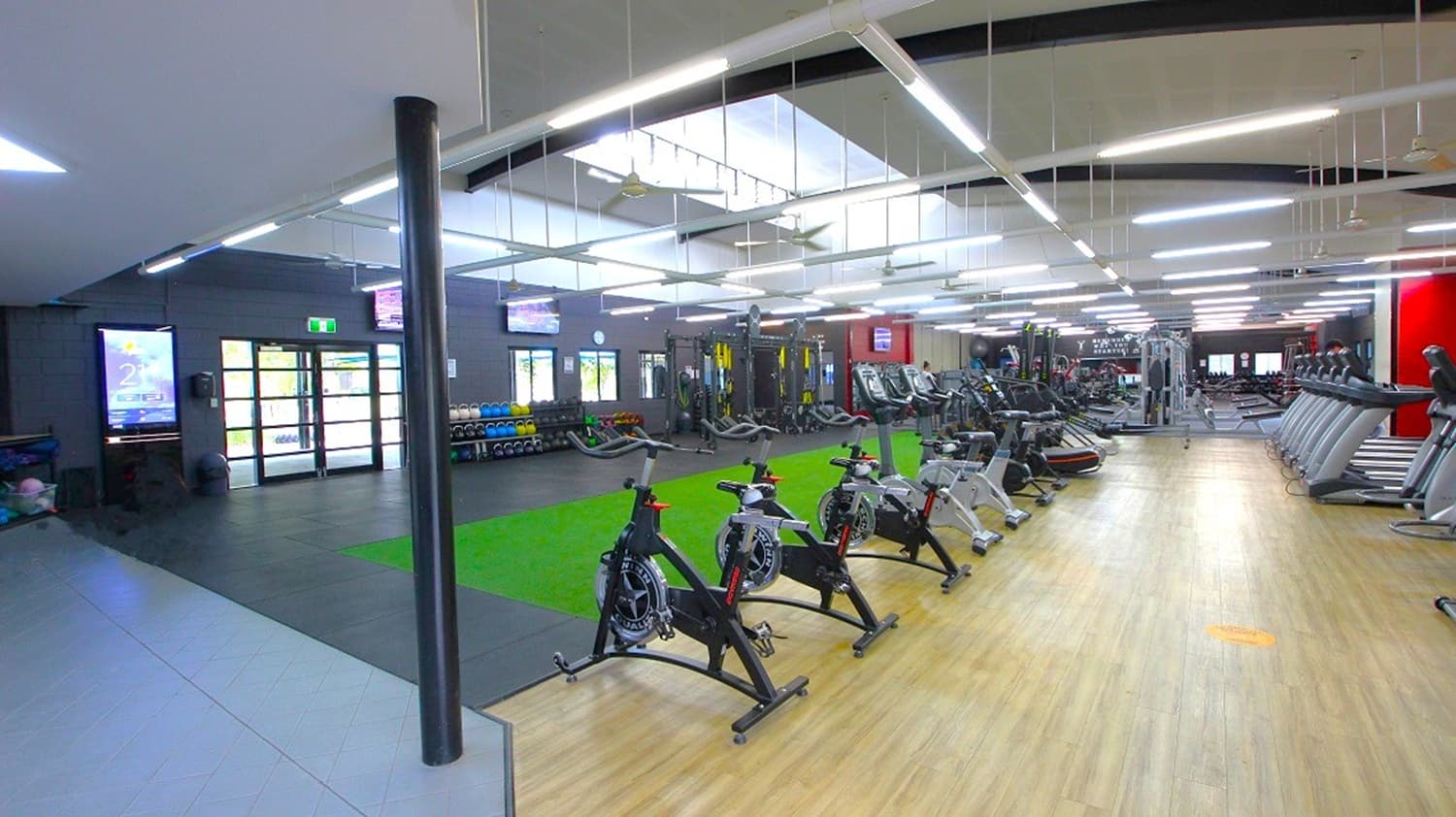 A spacious gym interior with a wooden floor and a high ceiling featuring linear lights. On the right, there are rows of treadmills and exercise bikes. In the centre, a green turf area is visible, surrounded by various gym equipment. The left side has a dark wall with large windows and a door, along with a rack of weights. The ceiling has skylights, allowing natural light to illuminate the space.