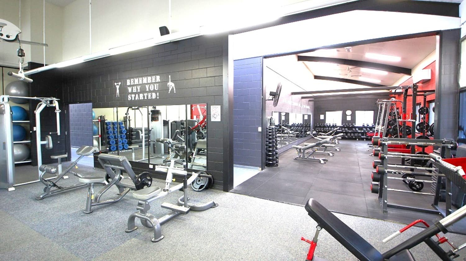 A gym interior with various exercise equipment. On the left, there are weight machines and benches on a grey carpeted floor. A black wall with a motivational sign reads "Remember Why You Started!" Mirrors reflect the equipment. On the right, more benches and weight racks are visible on a darker floor. The ceiling is high with exposed beams and bright lighting.