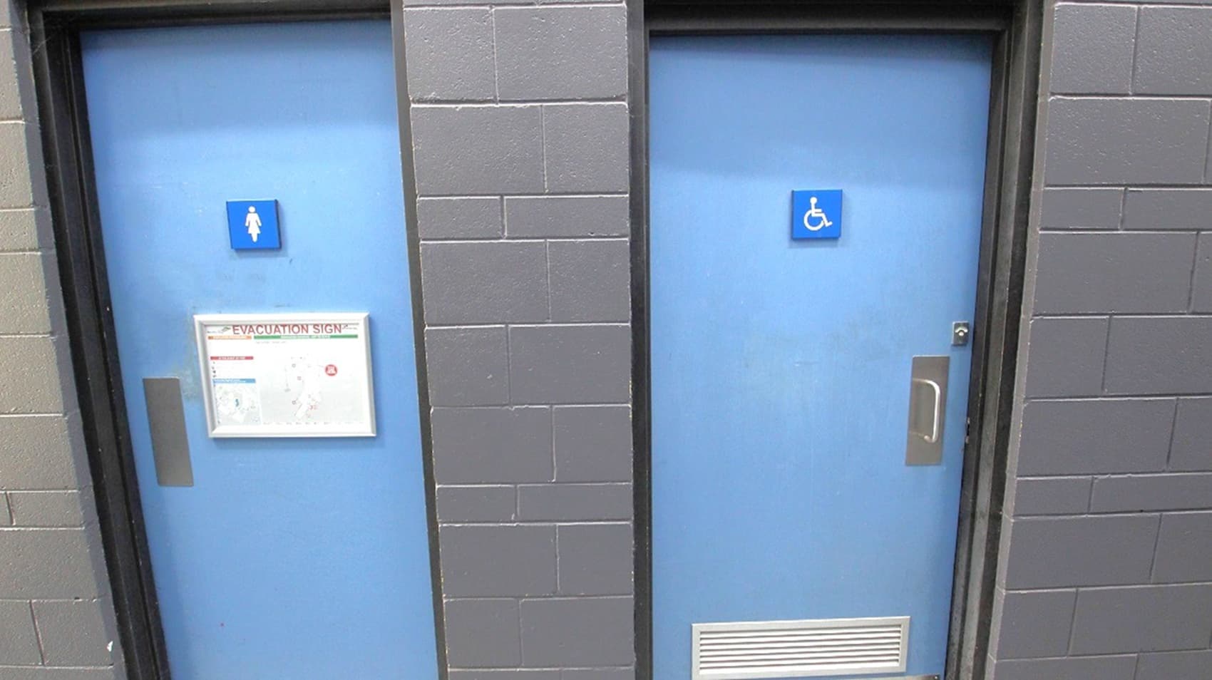 Two blue doors set in a grey brick wall. The left door has a sign indicating a women's restroom and a framed evacuation notice. The right door features a sign for accessible facilities and a vent at the bottom. Both doors have metal handles.