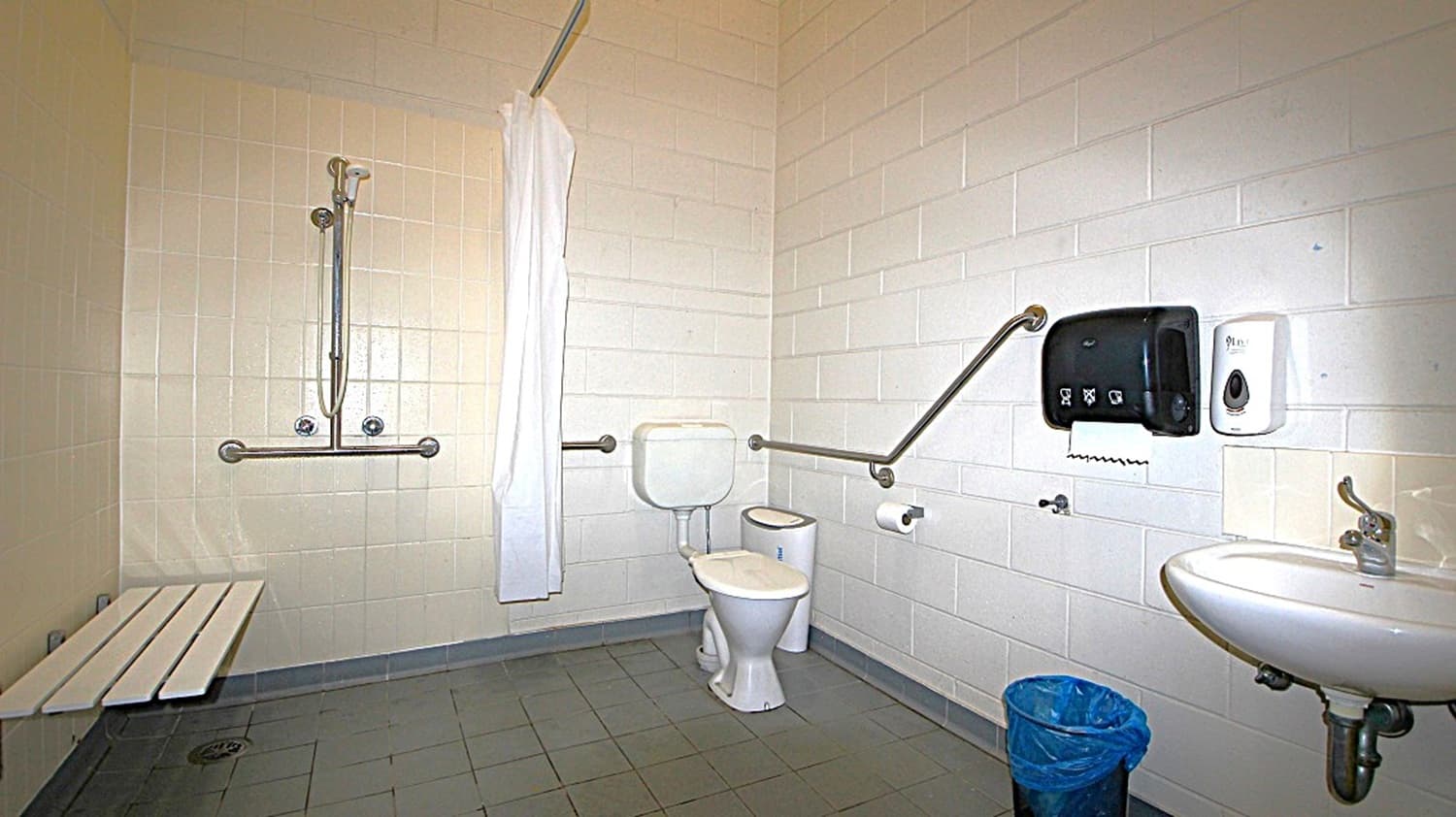 A bathroom with accessibility features. On the left, there's a shower area with a handheld showerhead and a white curtain, accompanied by a foldable bench. In the centre, a toilet with support rails on both sides. On the right, a wall-mounted sink with a tap, a paper towel dispenser, and a soap dispenser above. The floor is tiled in grey, and a blue bin is placed near the sink. The walls are tiled in white.