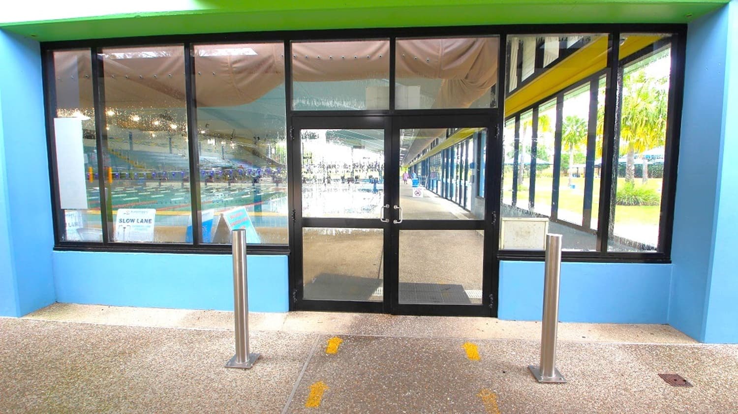 An entrance to an aquatic centre with blue walls and large black-framed windows on the left and right, flanking a central glass door. The foreground has a speckled beige floor with two stainless steel bollards, one on each side of the entrance. In the background, the interior gym area is visible through the left window, and a walkway with more glazing and yellow structural beams runs along the right.