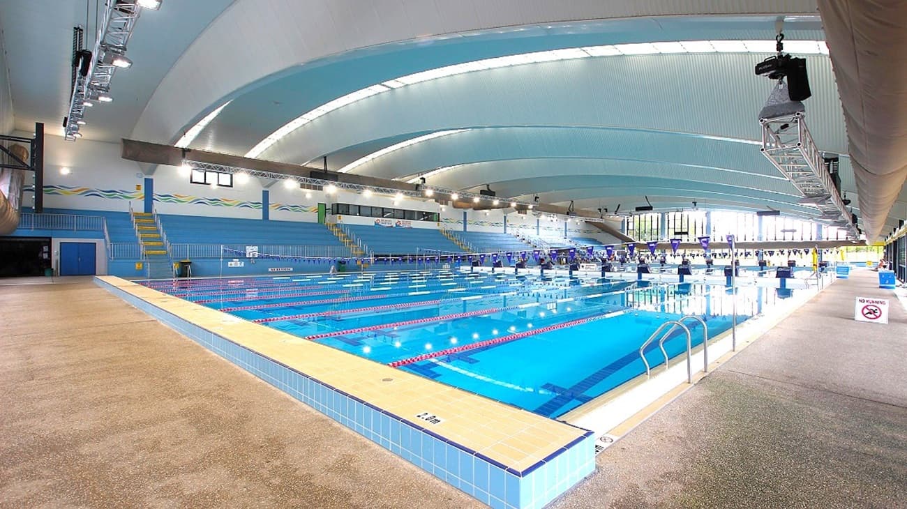 An indoor swimming pool with clear marked lanes occupies the middle, bordered by light beige textured flooring. To the left, blue seating tiers rise against the wall, with yellow railings. On the right, pool steps with metal handrails lead into the water. The background features large windows letting in light, while the high arched ceiling is pale blue with white sections and overhead lighting rigs.