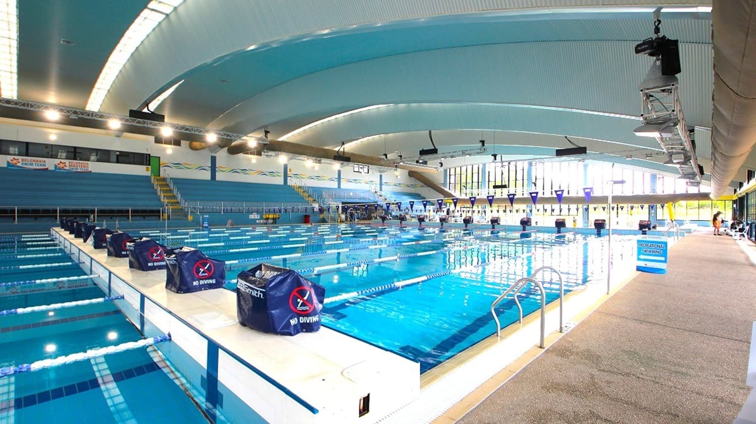 Blue-seated spectator stands with yellow railings are to the left, while the right features a wide beige textured walkway beside the pool. The middle holds a clear blue lane pool with marked starting blocks covered in "No Diving" covers. In the background, tall glass windows line the far wall. The curved ceiling above is pale blue with lighting panels and exposed fixtures.