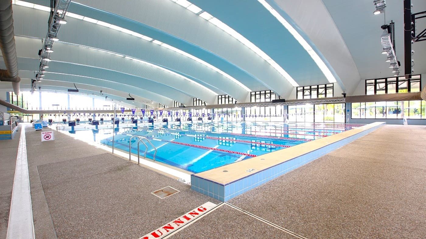 On the left, textured beige flooring runs beside a pool with metal handrails. The middle features a large tiled swimming pool with floating lane markers, while the right has more open floor space. The background is lined with tall glass windows. Overhead, the ceiling is curved with alternating blue and white panels and evenly spaced lighting fixtures.
