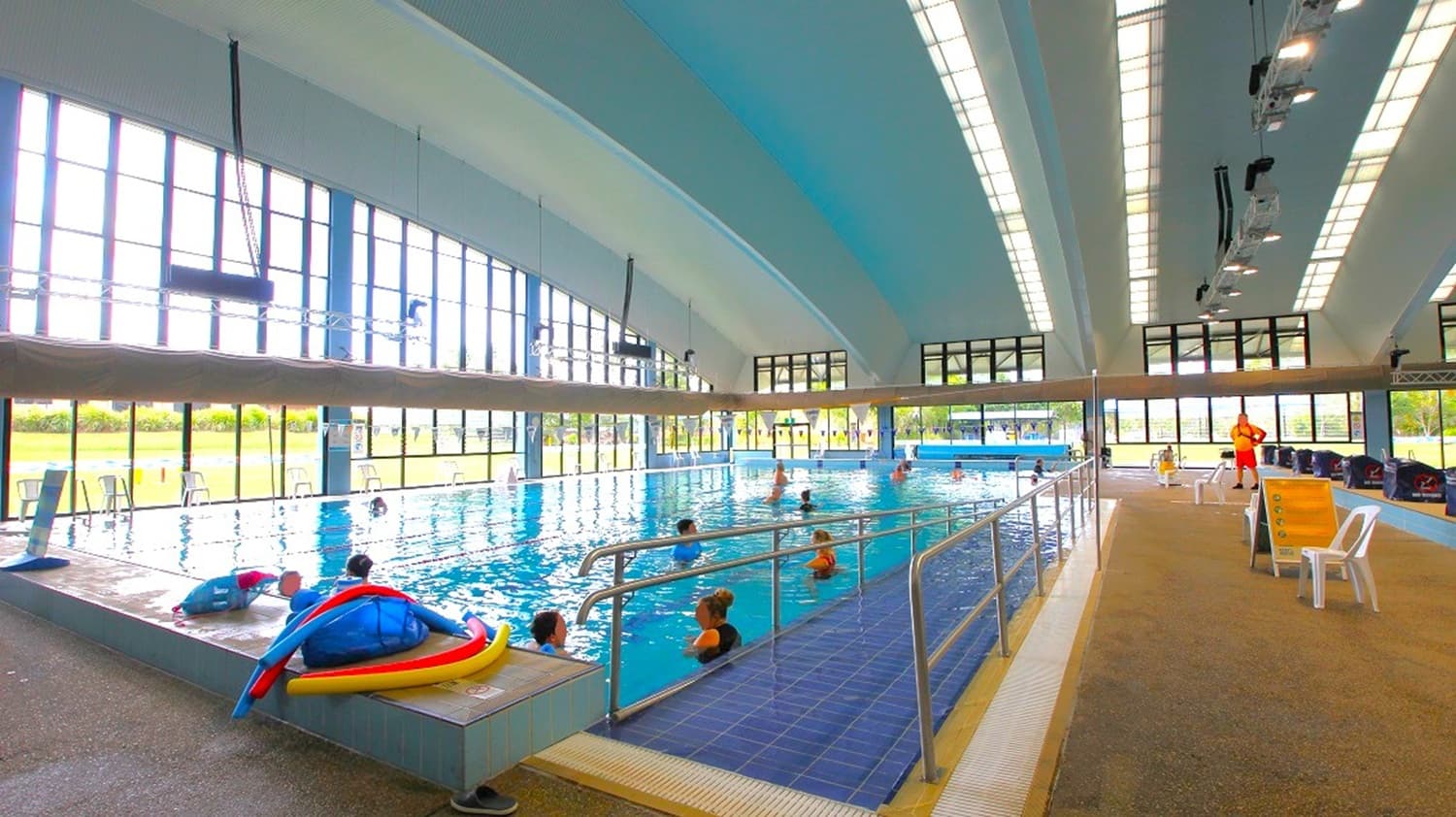 On the left, poolside equipment and colourful floatation aids sit at the pool’s edge. The middle contains a spacious swimming pool with a ramp and handrail, where people are swimming. The right features beige textured flooring with white plastic chairs and a lifeguard. The background wall consists of tall windows, and the ceiling is high with blue and white curved panels and overhead lighting.