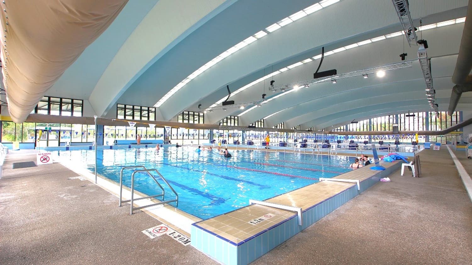 On the left, a large indoor air duct runs above beige textured flooring, with a poolside metal handrail. The middle features a blue-tiled swimming pool with people in the water, steps leading in, and lane ropes. On the right, there are white plastic chairs and pool toys near the edge. The background is lined with tall windows, and above, a curved, blue-and-white panelled ceiling with exposed lighting and equipment.